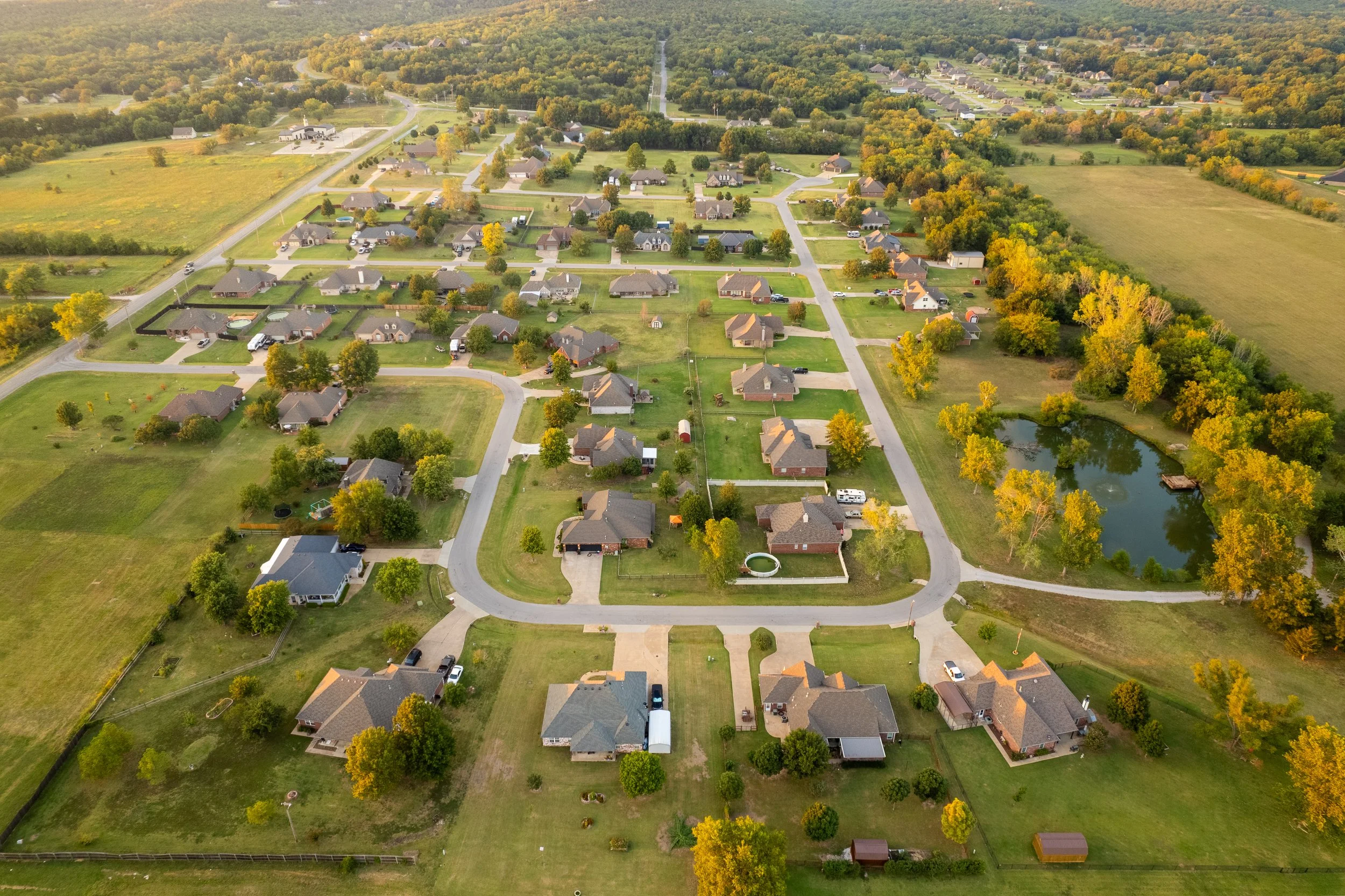 Aerial view of a suburban neighborhood with houses, green lawns, trees, and a pond surrounded by trees.