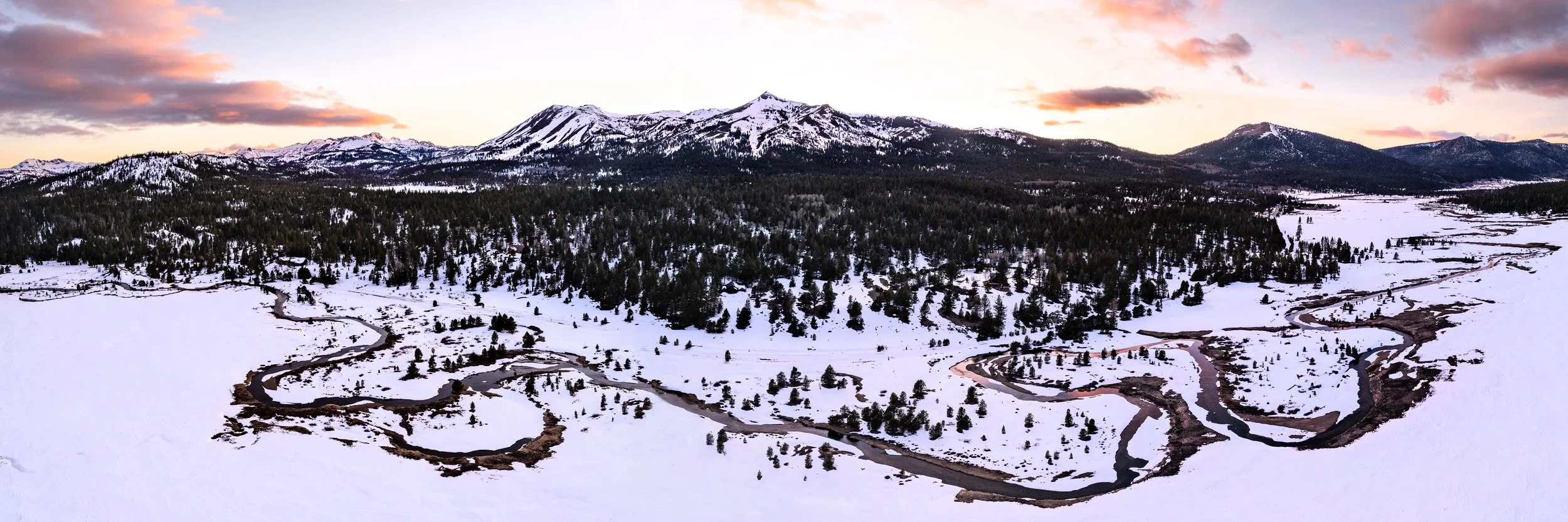 Snow-covered valley with winding rivers, dense forest at the base of snow-capped mountains, and a colorful sunset sky with pink and purple clouds.