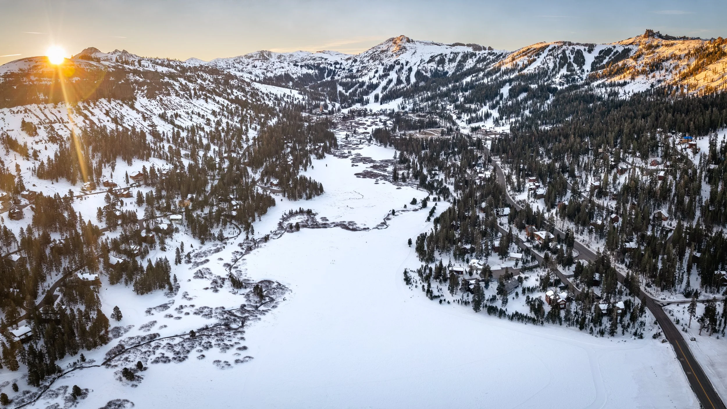 Snow-covered mountain landscape at sunrise with a frozen lake, trees, and houses along a winding road.