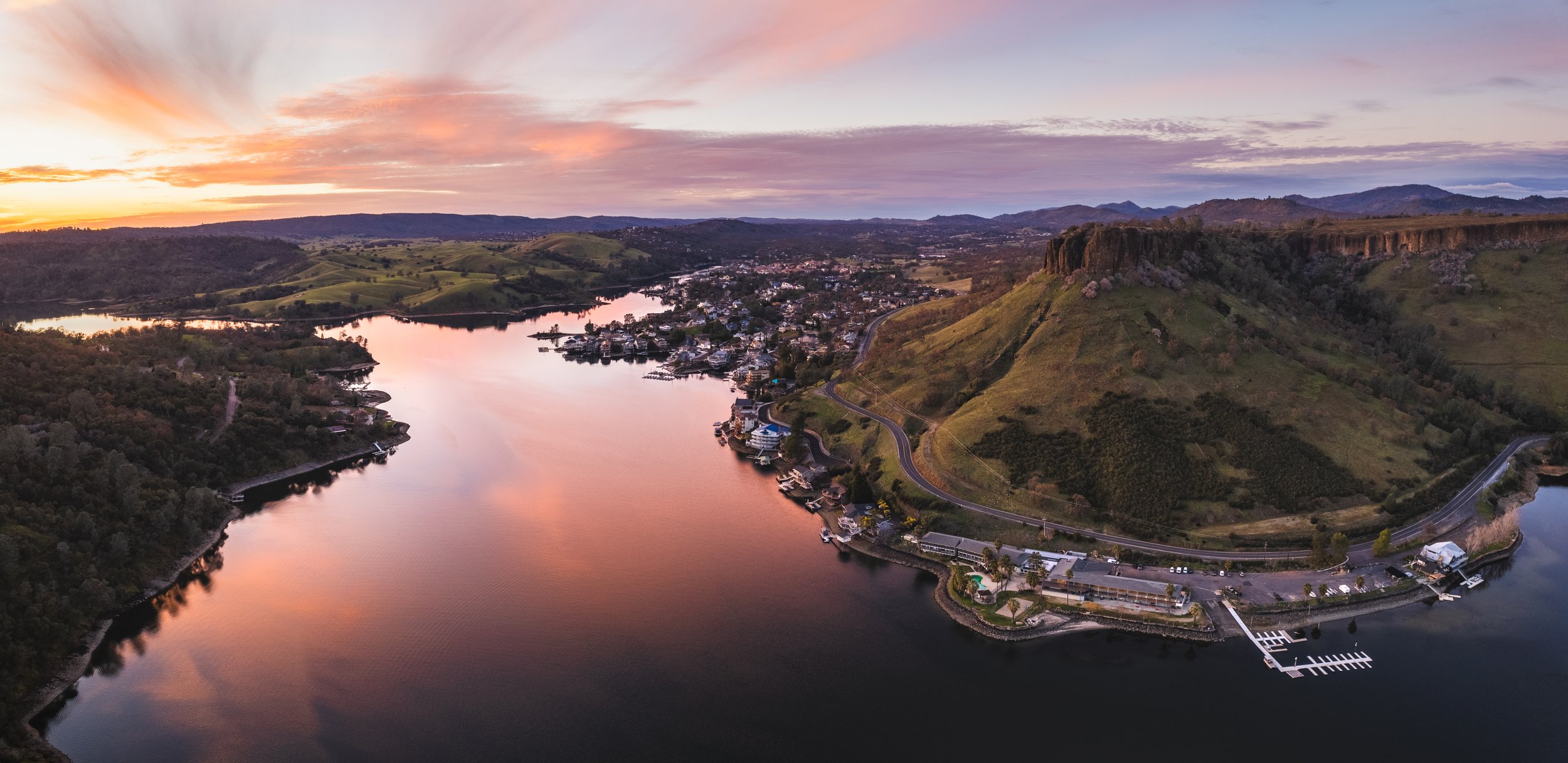 Aerial view of a river at sunset with surrounding green hills, a small town on the riverbank, and a marina with boats near a shoreline