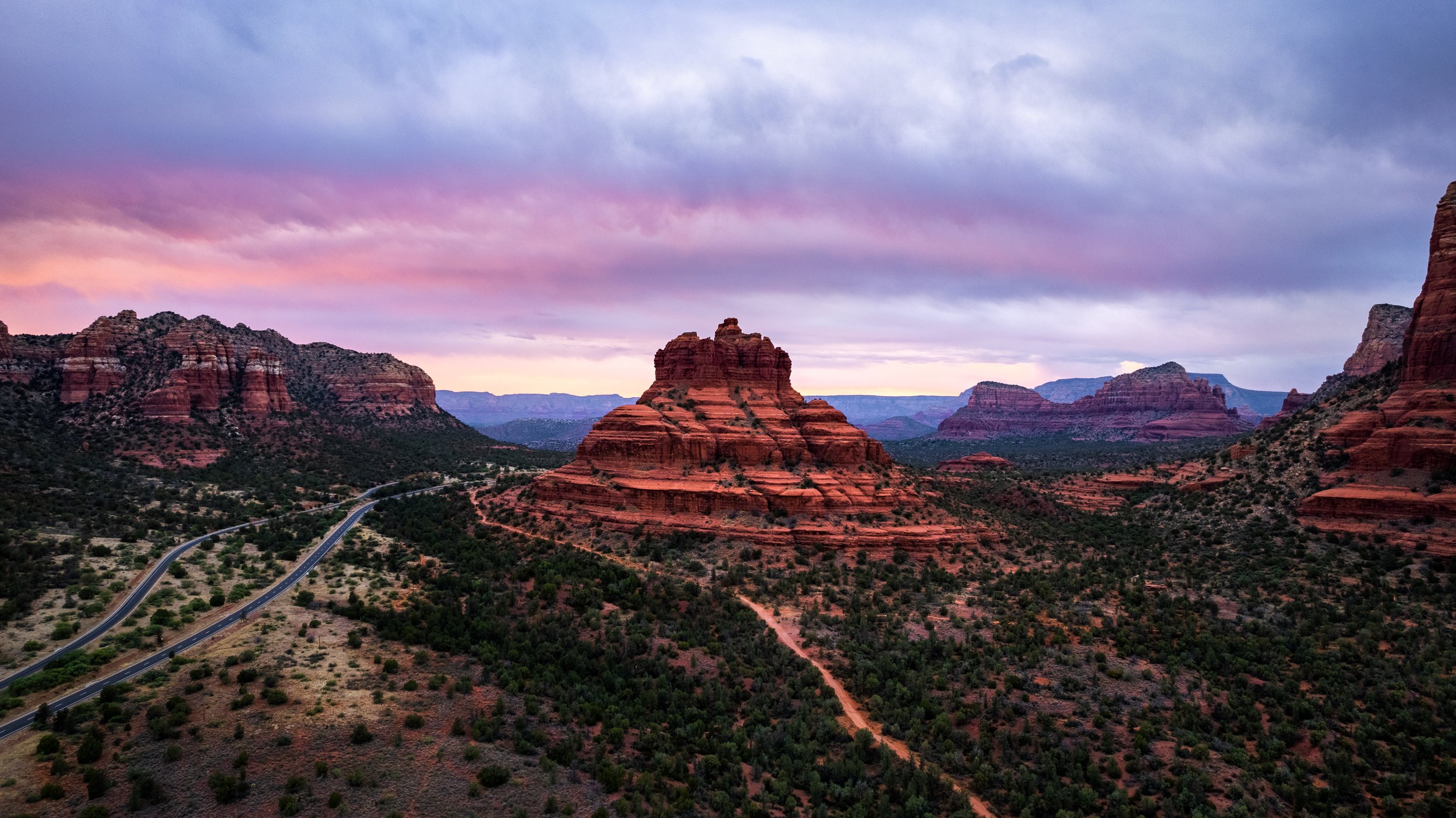A landscape of red rock formations and mesas in a desert, with a winding road in the foreground and a colorful cloudy sky overhead.
