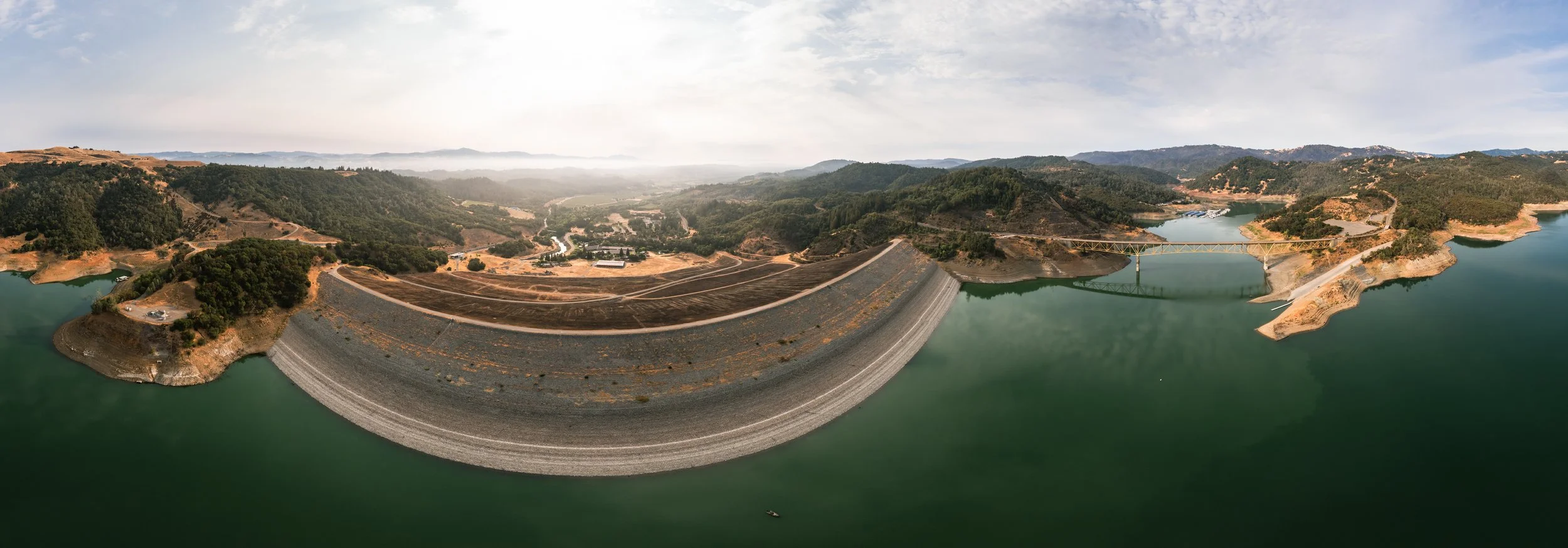 Aerial view of a large dam on a river with surrounding hills and water conservation areas. The dam is curved and holds back a reservoir with green water, with a bridge connecting the two sides of the reservoir.