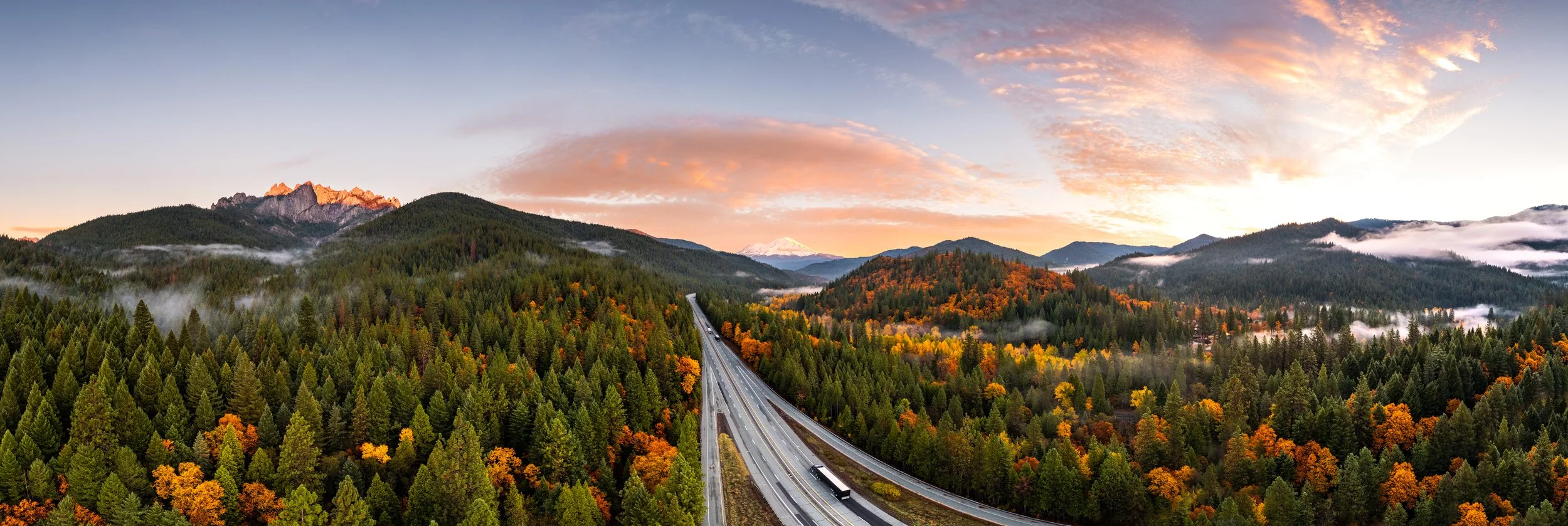 A scenic landscape of a highway winding through a forested valley with colorful autumn trees, rolling hills, mountains, and a partly cloudy sky at sunset.