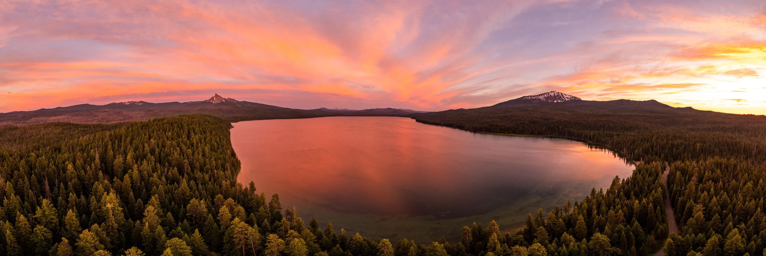 A panoramic view of a large lake surrounded by dense forest with mountains and snow-capped peaks in the distance, under a colorful sunset sky with pink, orange, and purple clouds.