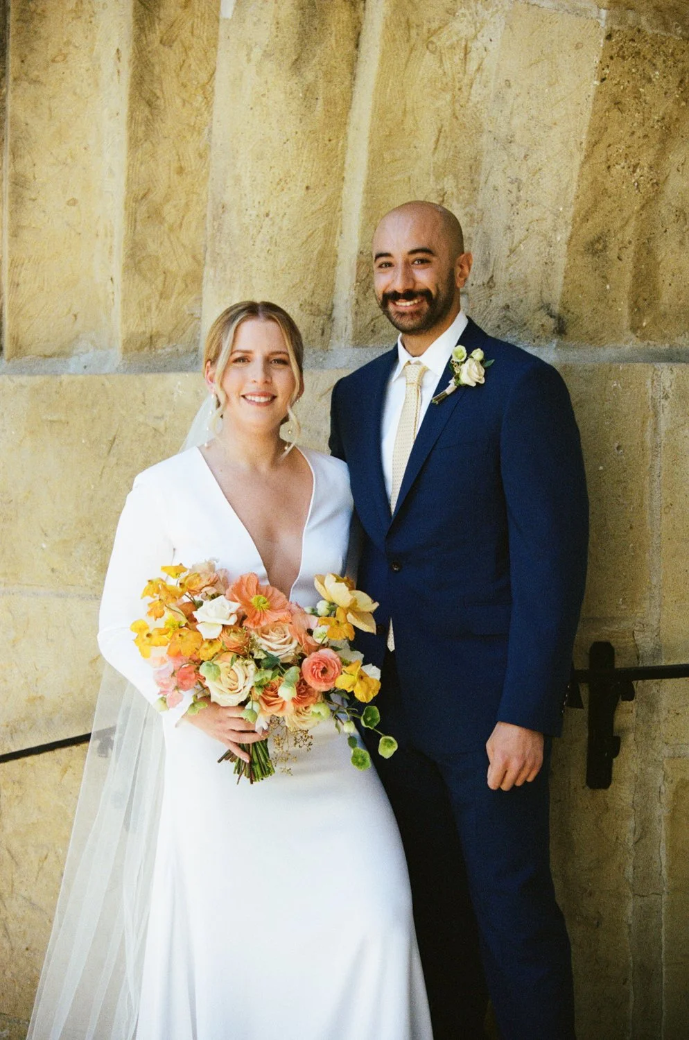 Modern house with wooden exterior surrounded by yellow flowers, mountains in the background, and a large glass window showing two people inside.