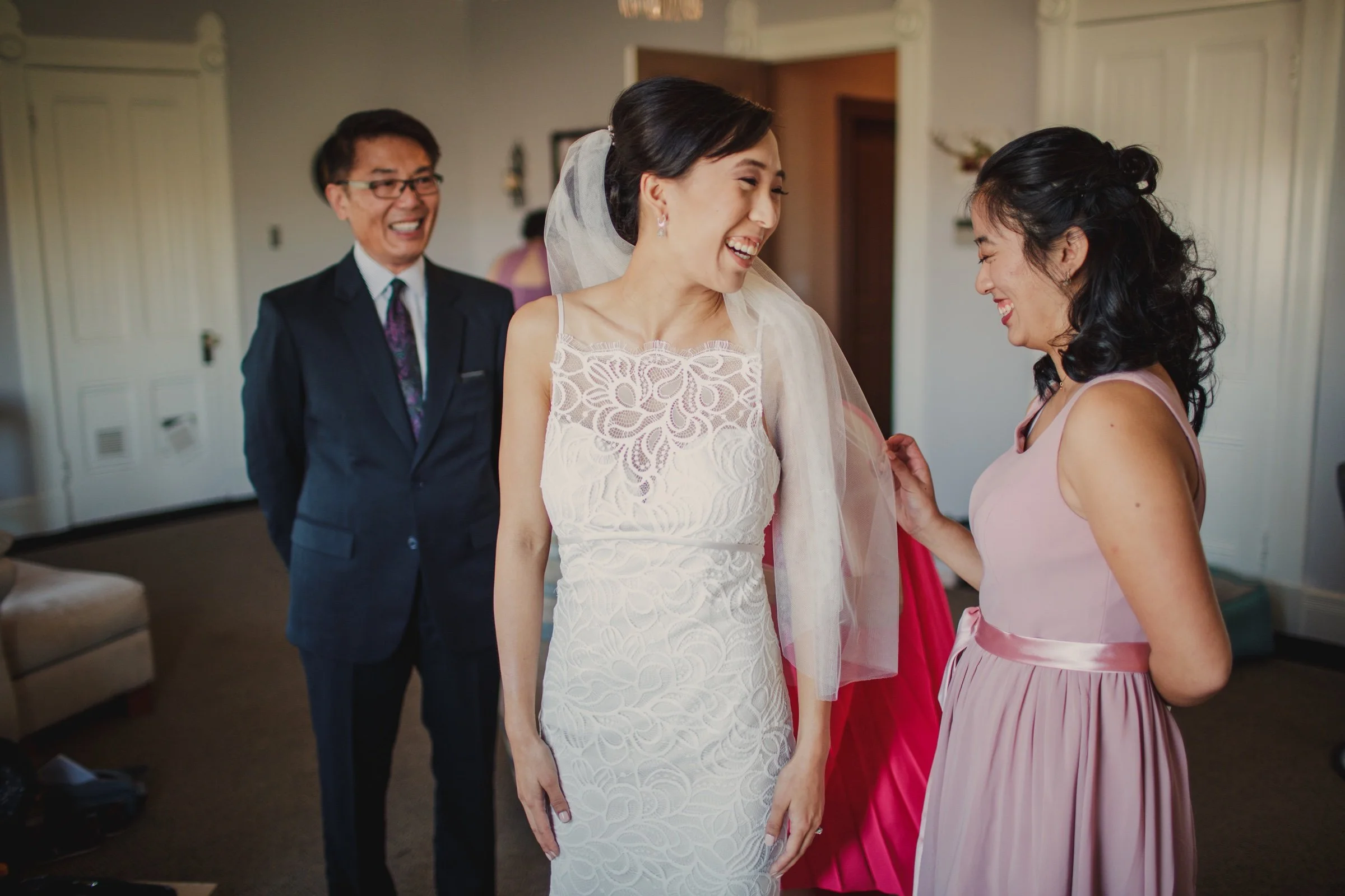 Bride laughing with a woman in a pink dress while a man smiles nearby.