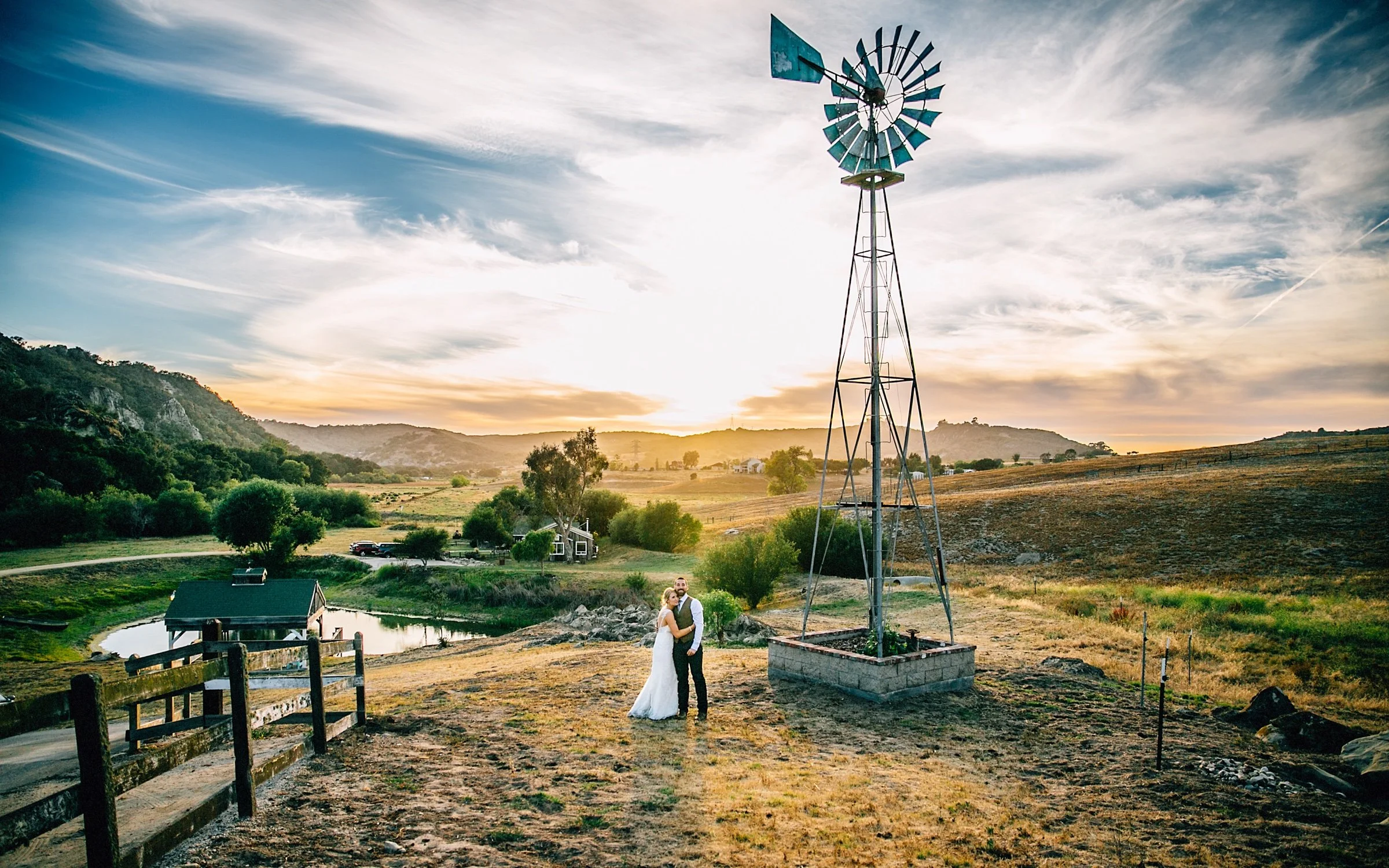Bride and groom beside a windmill in rolling hills at sunset.