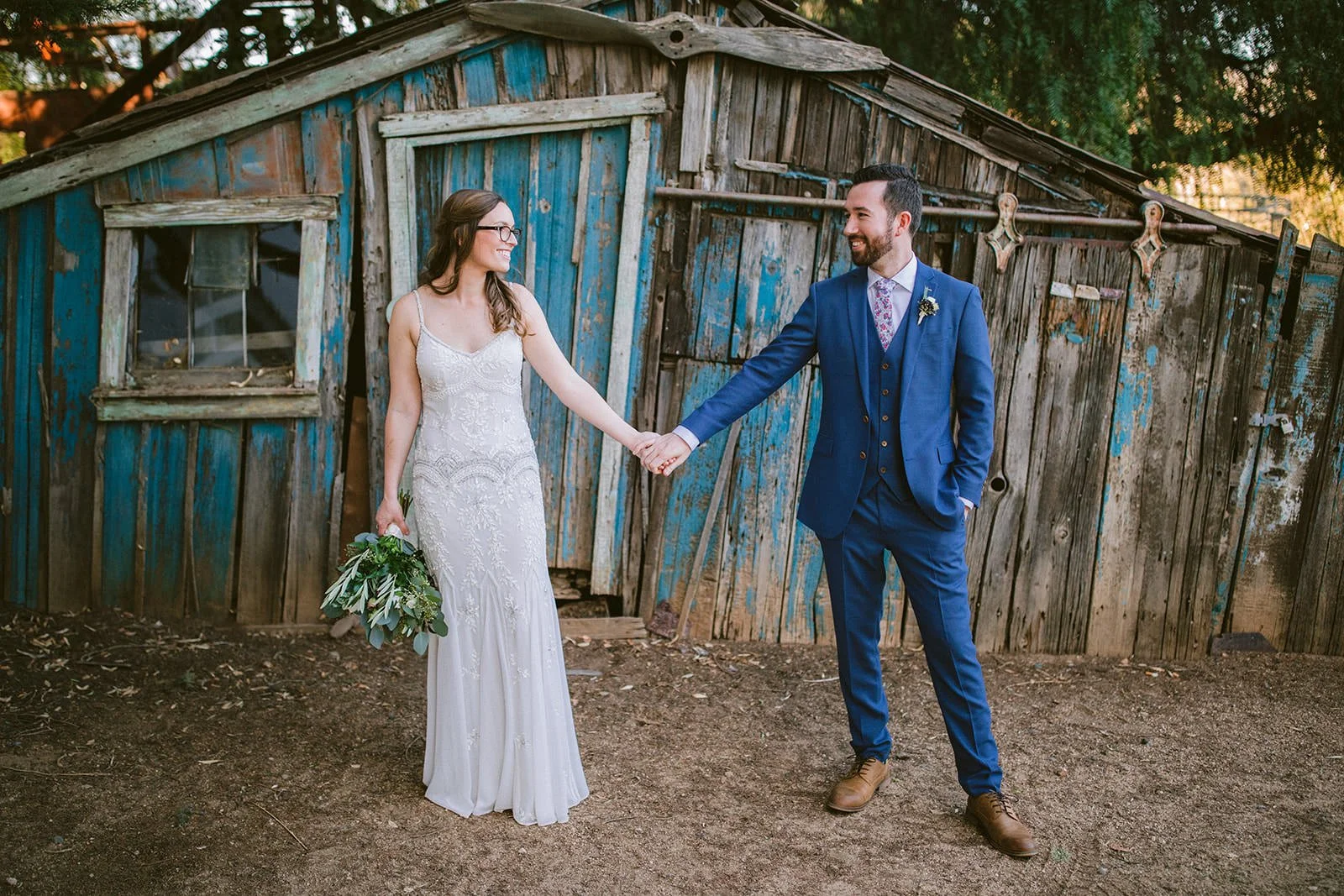 Bride and groom holding hands in front of weathered blue barn shed at Flying Caballos Ranch, Central Coast wedding photography
