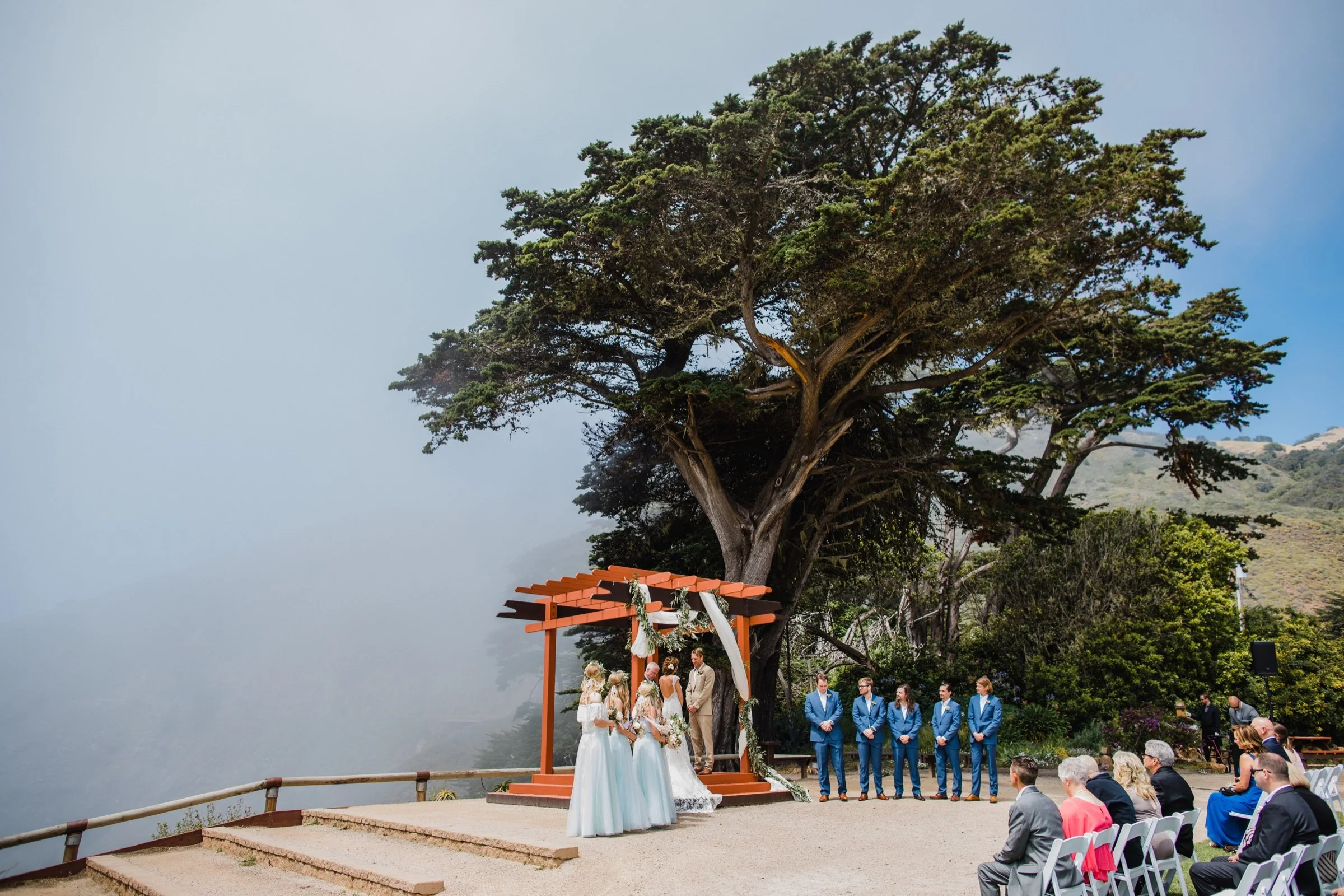 Coastal wedding ceremony beneath a large tree beside a wooden arbor.