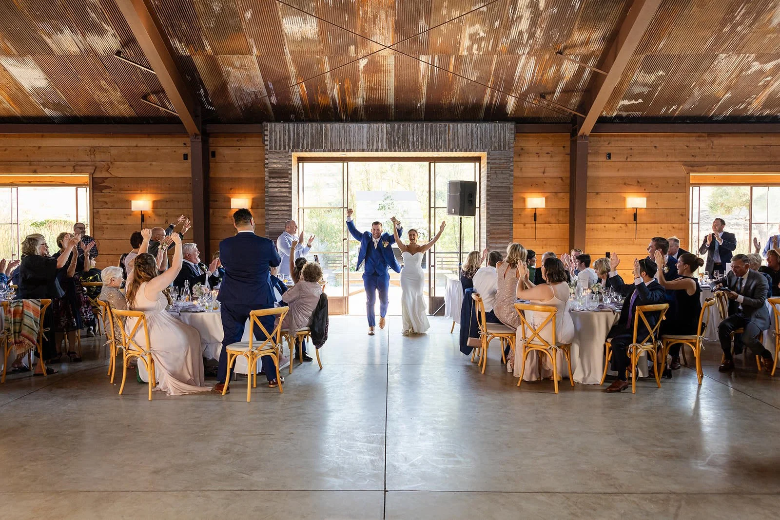 Bride and groom grand entrance into Flying Caballos Ranch Barn reception, arms raised, guests cheering