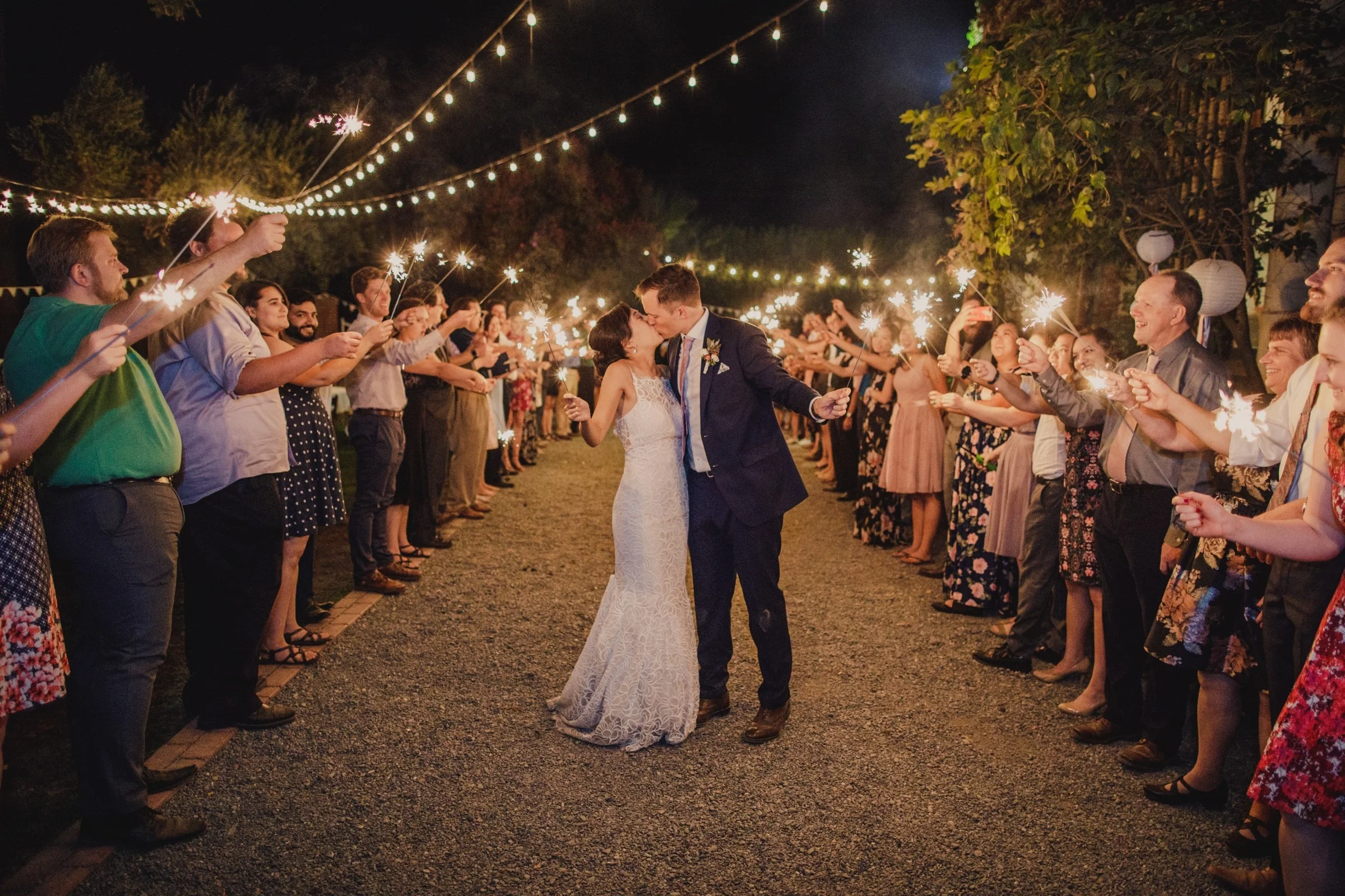 Bride and groom kissing as guests hold sparklers beneath string lights.