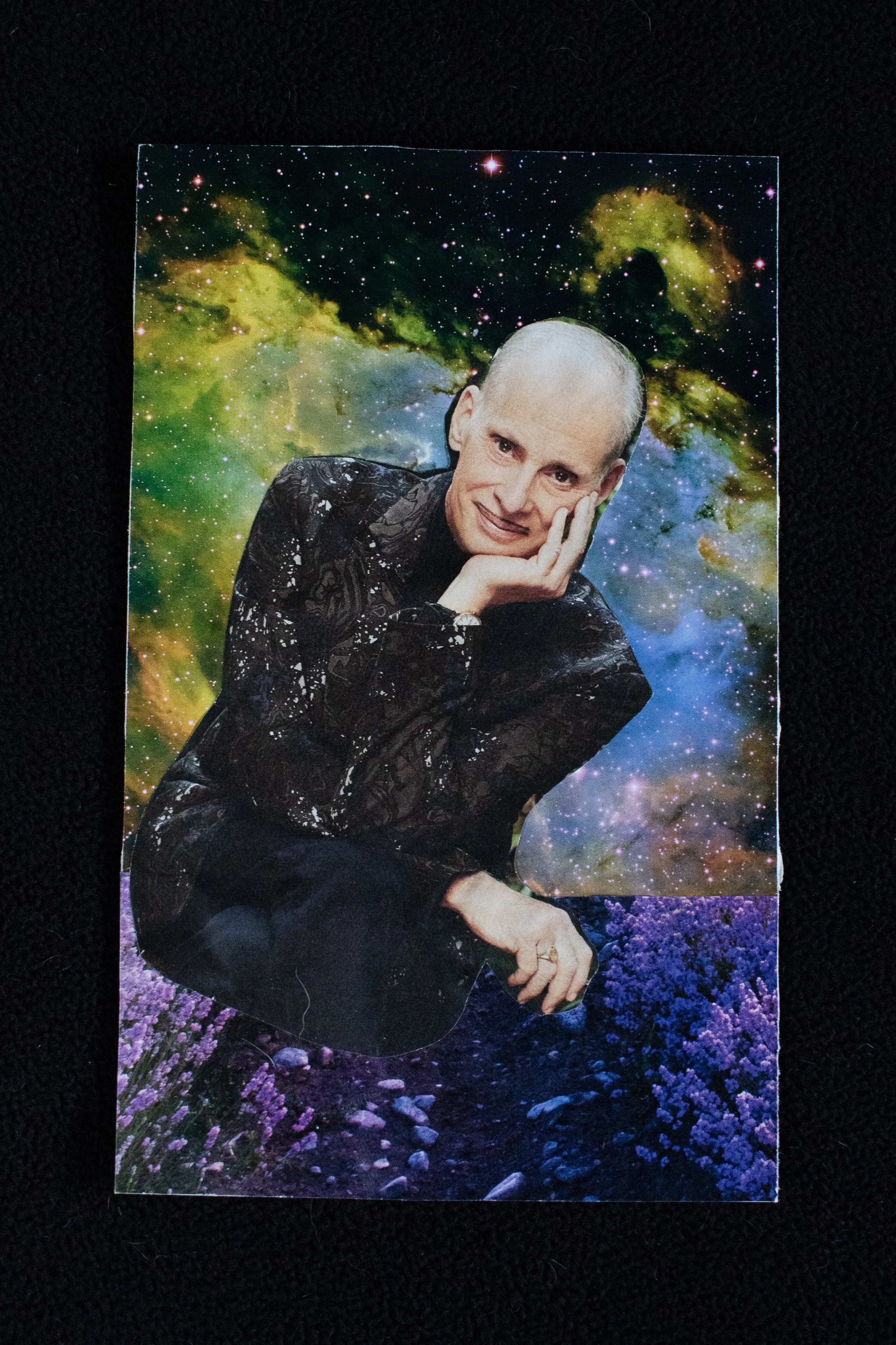 A collage of a person with a bald head, smiling, resting their chin on their hand, with a galaxy background and purple flowers on the ground.