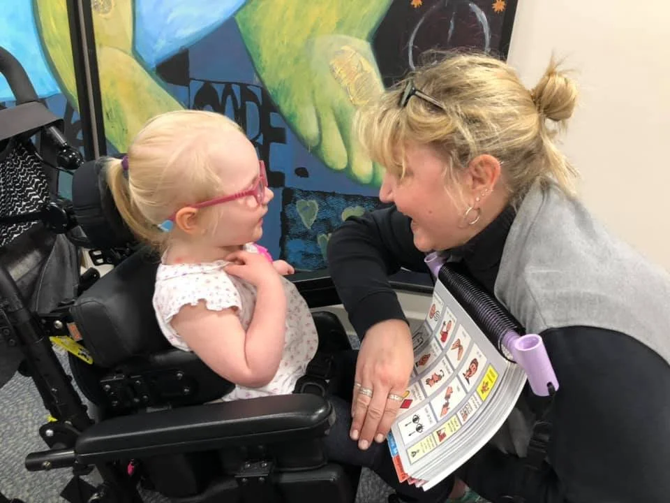 A woman leaning over a girl with cerebral palsy, sitting in a wheelchair, having a close, smiling conversation. They are indoors near a colorful mural.