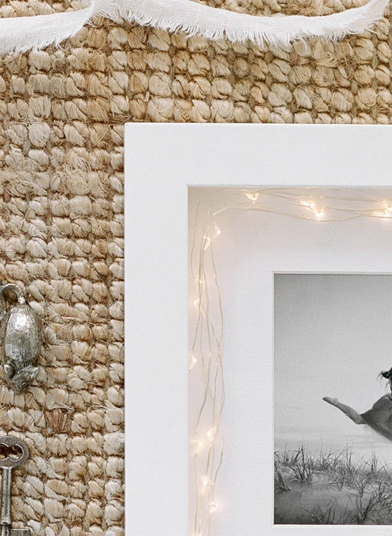 Close-up of a decorated wall corner showing a textured, woven beige surface, a white picture frame with string lights, and part of a black and white photo inside the frame.