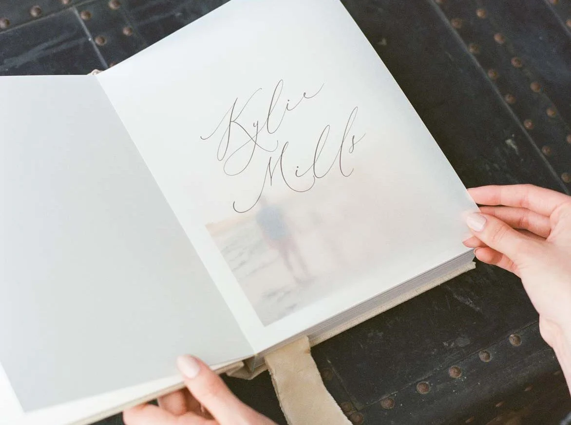 Open family album with handwritten names "Kylie" on the page, held by a person's hands over a dark surface.
