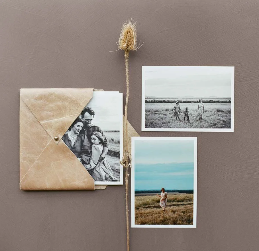 A brown leather envelope with black and white photographs inside, alongside two color photographs of a woman walking in a field, and a dried flower on a plain background.