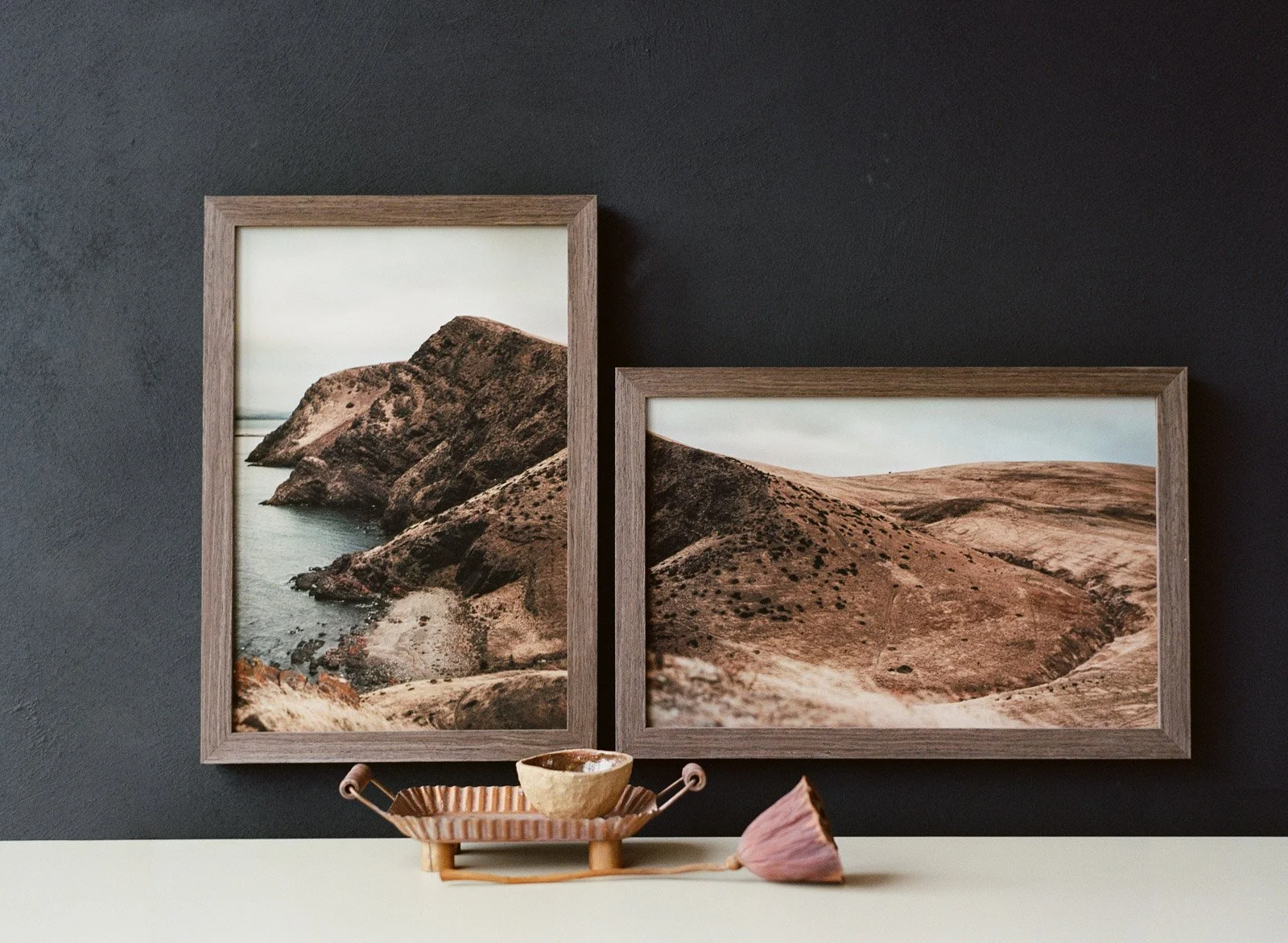 Two framed landscape photographs of rocky hills and coastline hang on a black wall, with a decorative shelf holding a bowl, a pink flower, and a small dish in the foreground at Atkins Photo Lab.