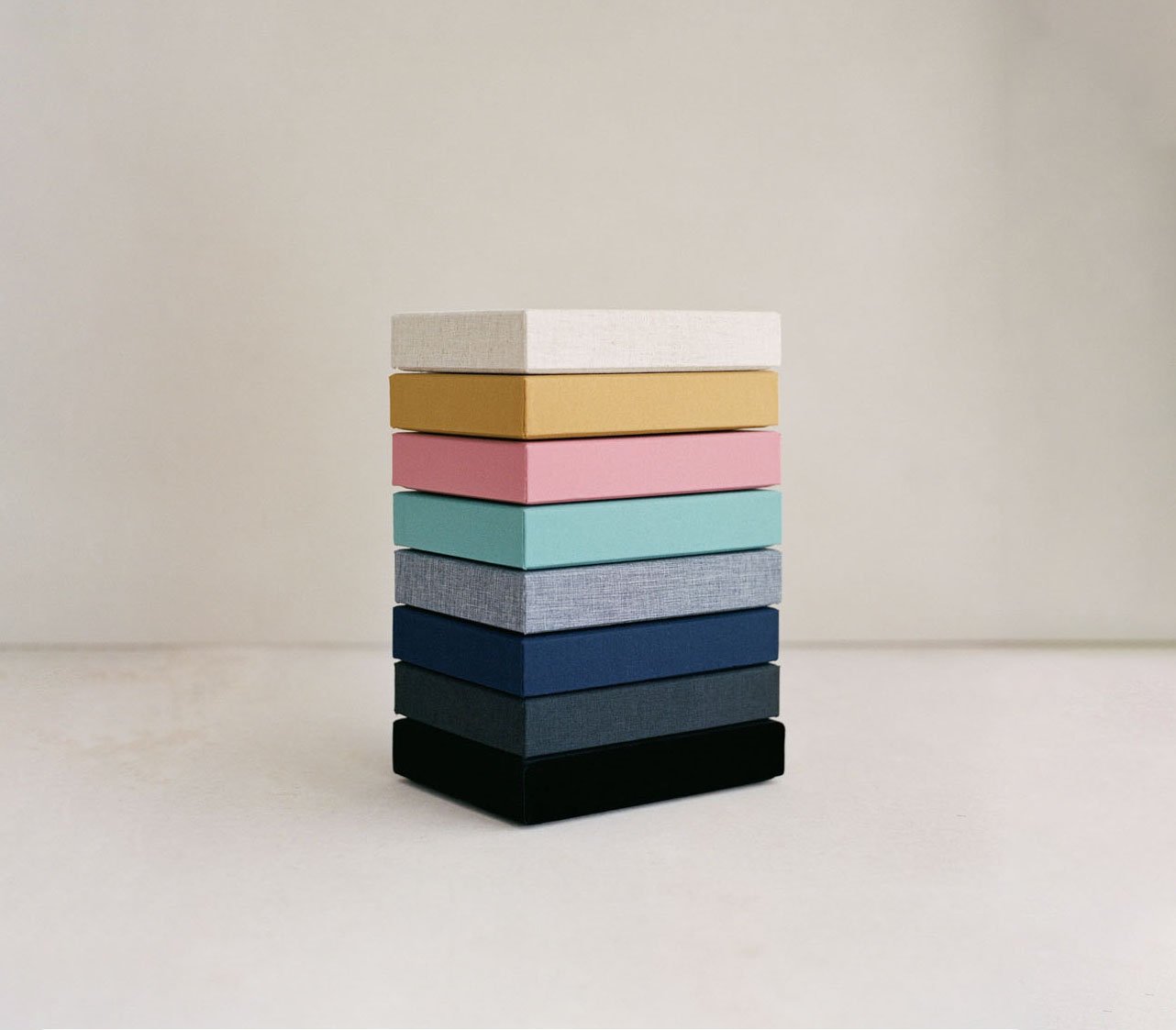 Stack of eight fabric-covered storage boxes in white, tan, pink, turquoise, gray, navy, dark gray, and black on a light-colored surface against a plain beige wall.