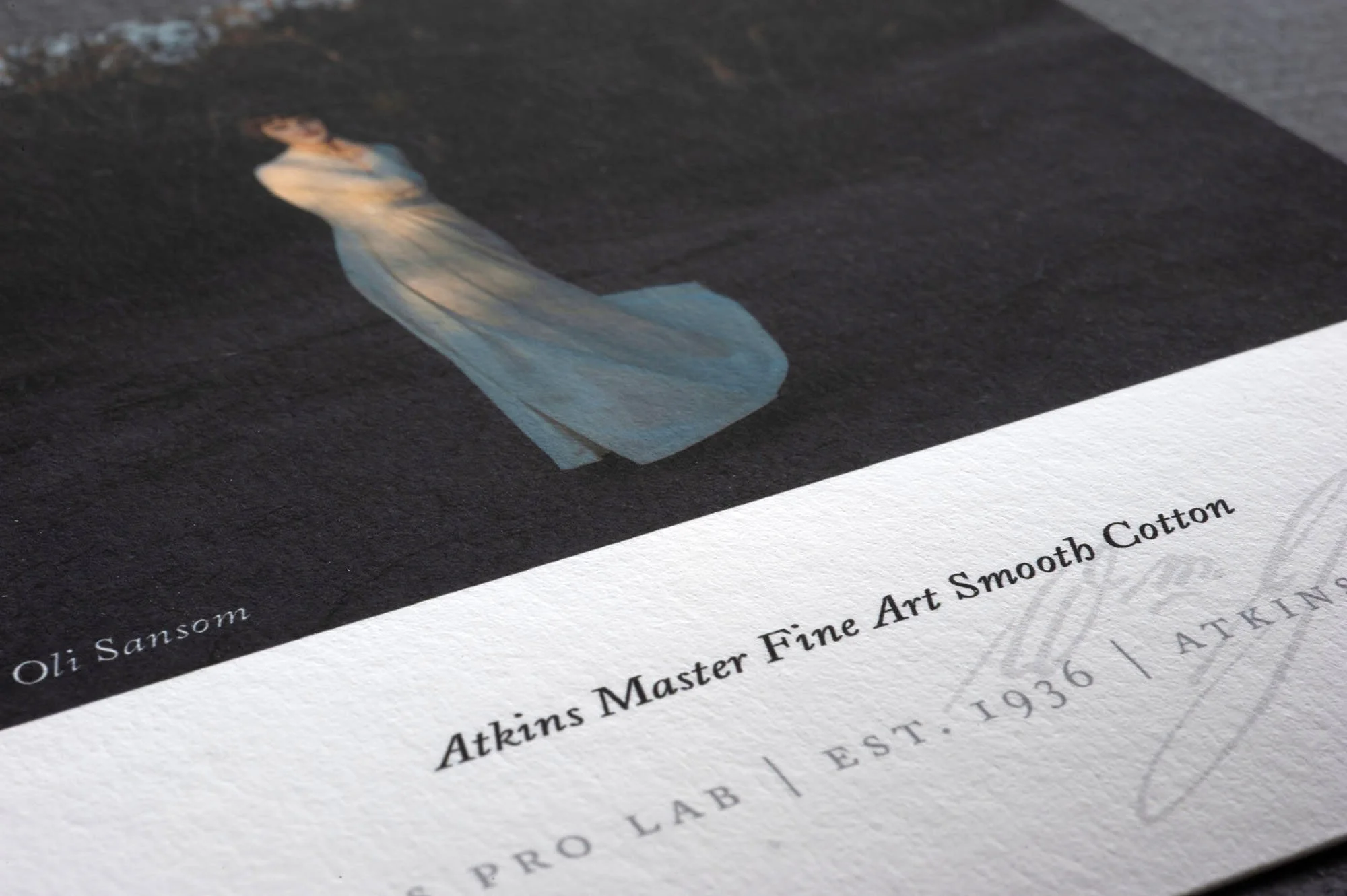 Close-up photo of a printed fine art photo print of a woman in a blue dress, with text indicating the artwork is by Oli Sansom and from the Atkins Master Fine Art Smooth Cotton paper at Atkins Photo Lab.