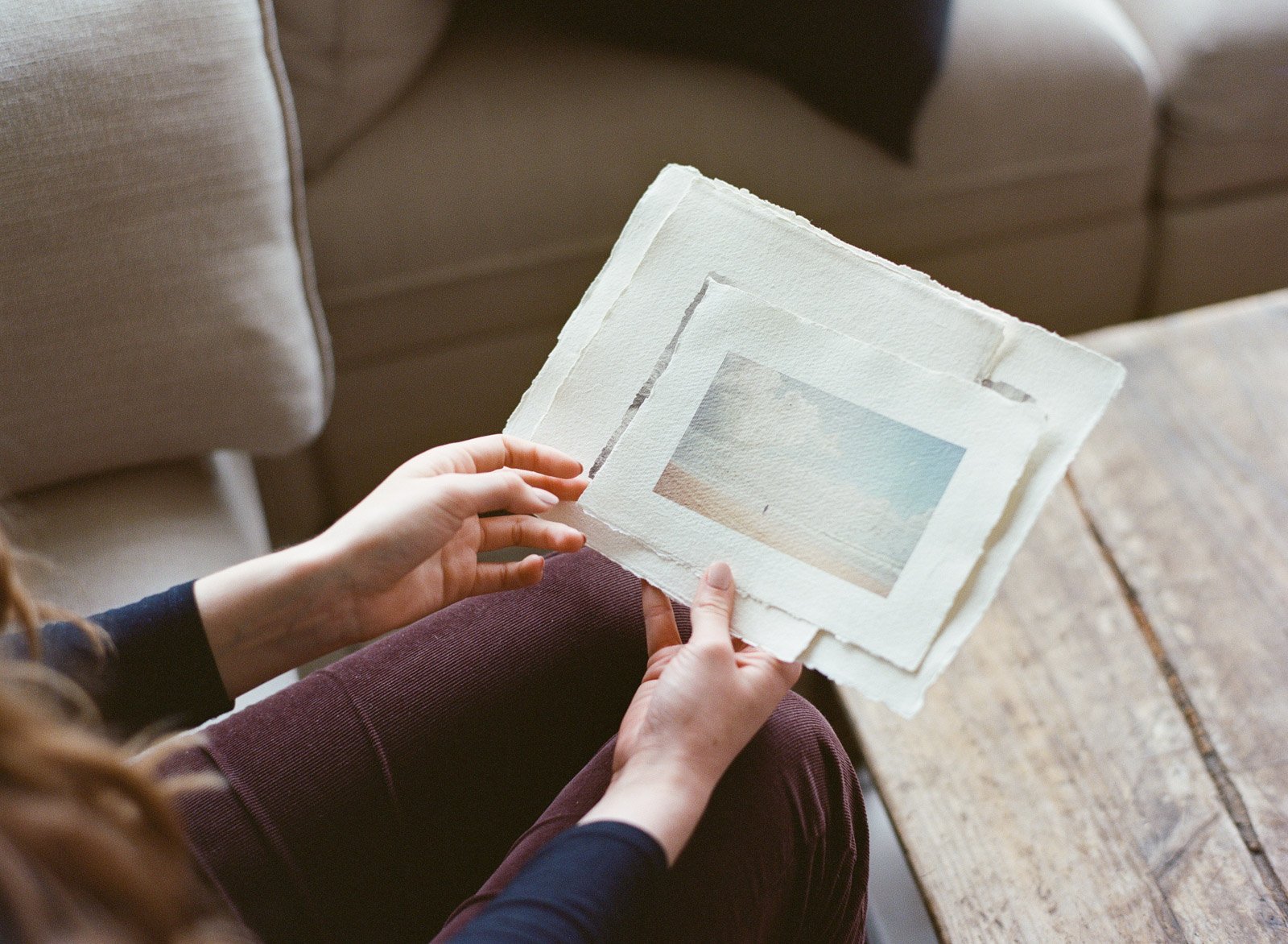 Person holding three watercolor landscape paintings on textured paper, sitting on a rustic wooden table.