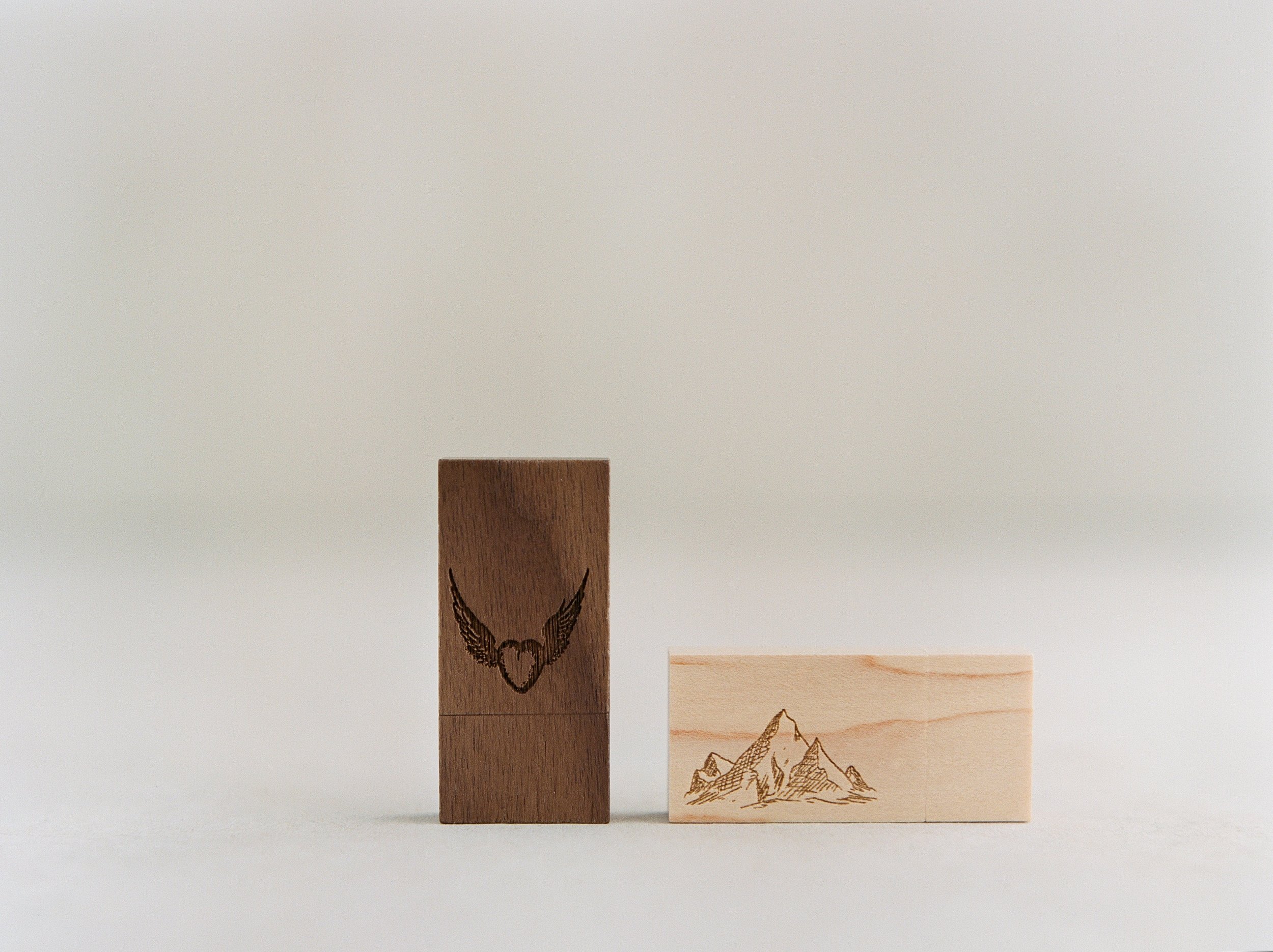 Two wooden usbs, one with a heart and wings design and the other with a mountain scene, on a plain background.