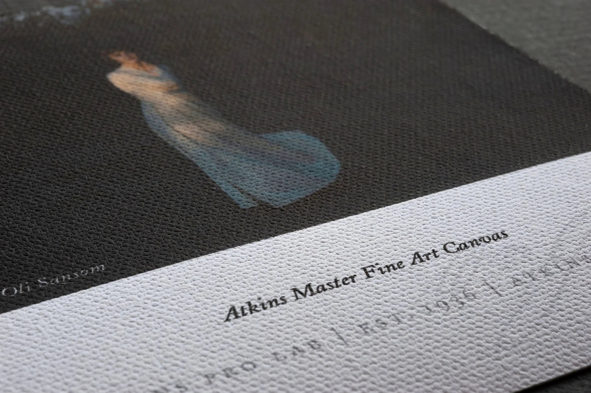 Close-up of a textured art gallery label with the text "Atkims Master Fine Art Canvas" and part of a painting of a woman lying on her side with brown hair, on a black background.
