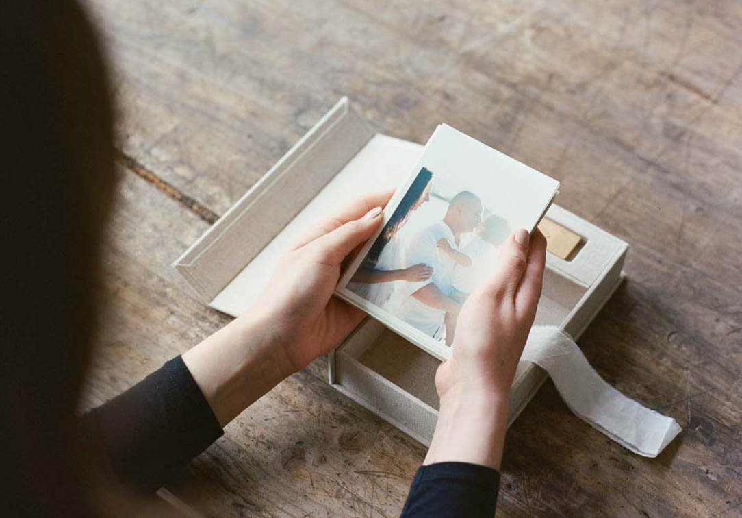 Person holding a photo album featuring a picture of a couple embracing, with the album placed inside a box on a wooden surface.