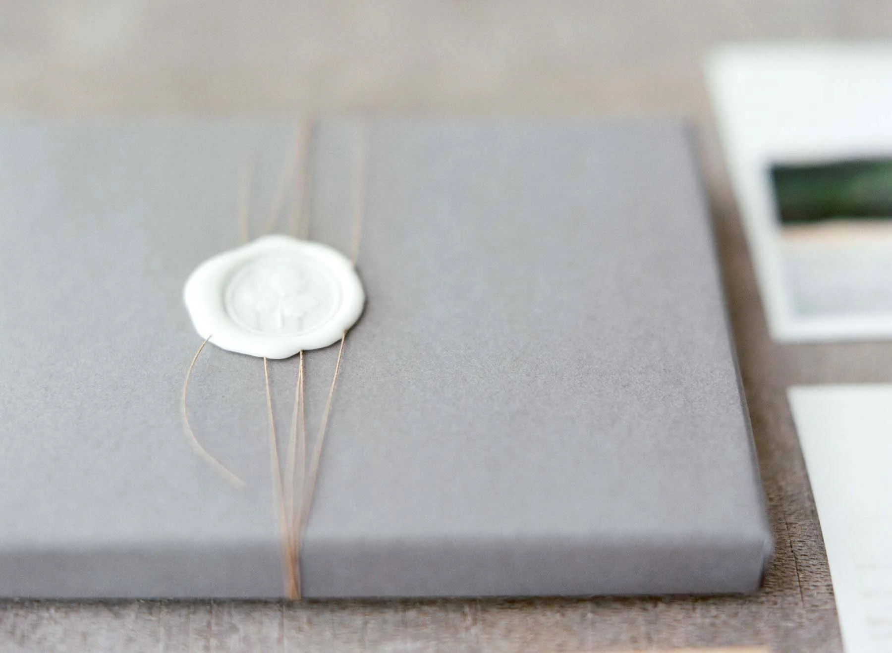 Gray gift box wrapped with a thin beige ribbon, sealed with a white wax seal with a decorative emblem.