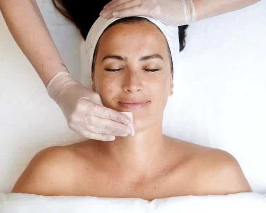 A woman receives a facial treatment from a professional wearing gloves, lying down with a relaxed expression.