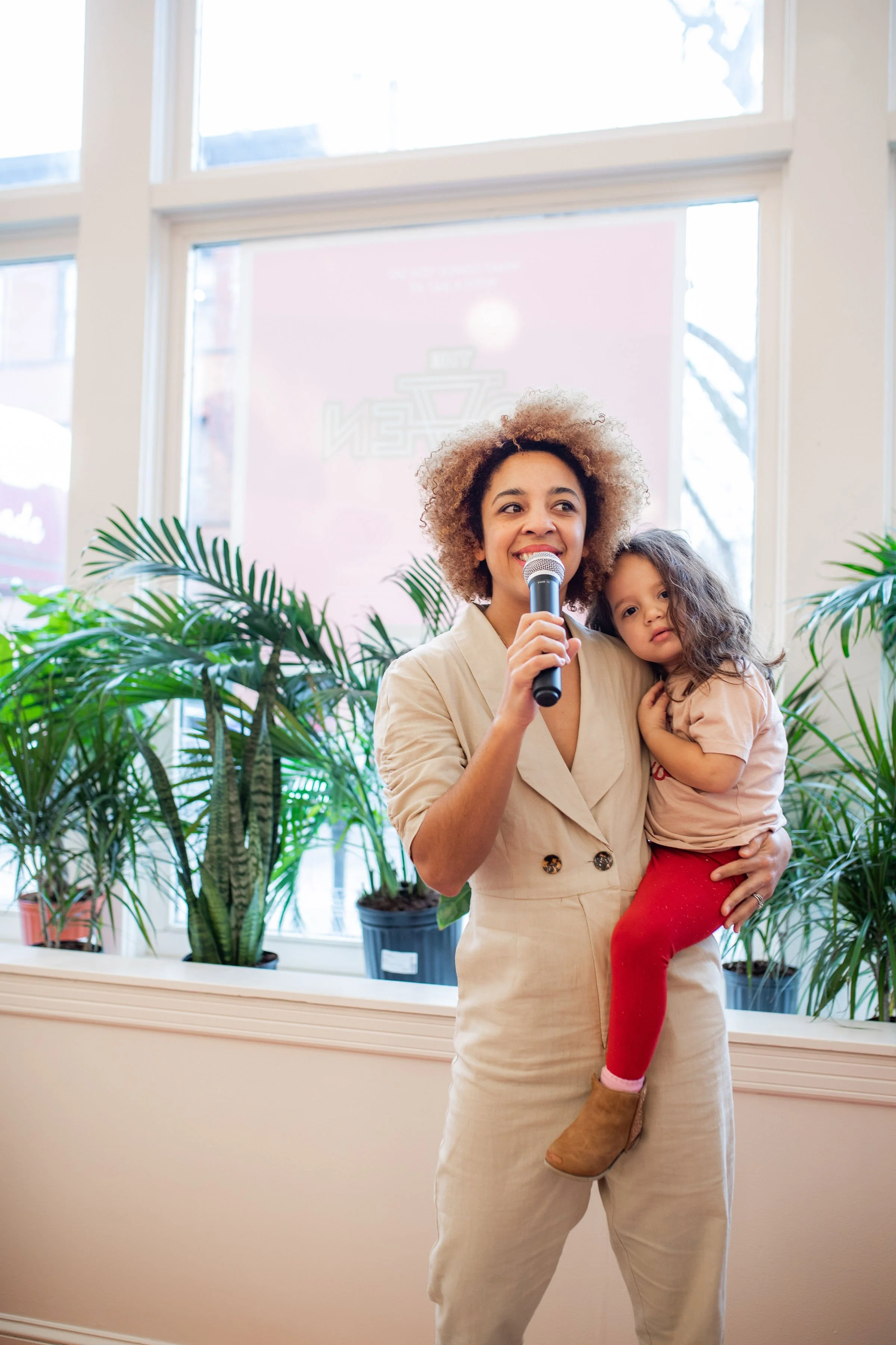 A woman holding a microphone and a young girl in her arms, standing in front of large windows with green potted plants, indoors during daytime.