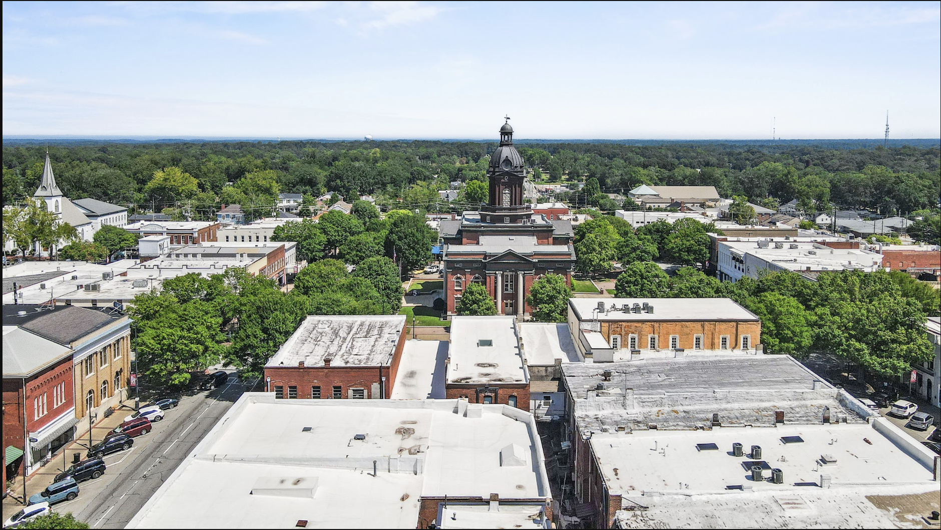 Aerial view of a small town with a prominent historic courthouse featuring a black dome and a cupola, surrounded by green trees and a mix of old and modern buildings, with a dense forest in the background under a clear blue sky.