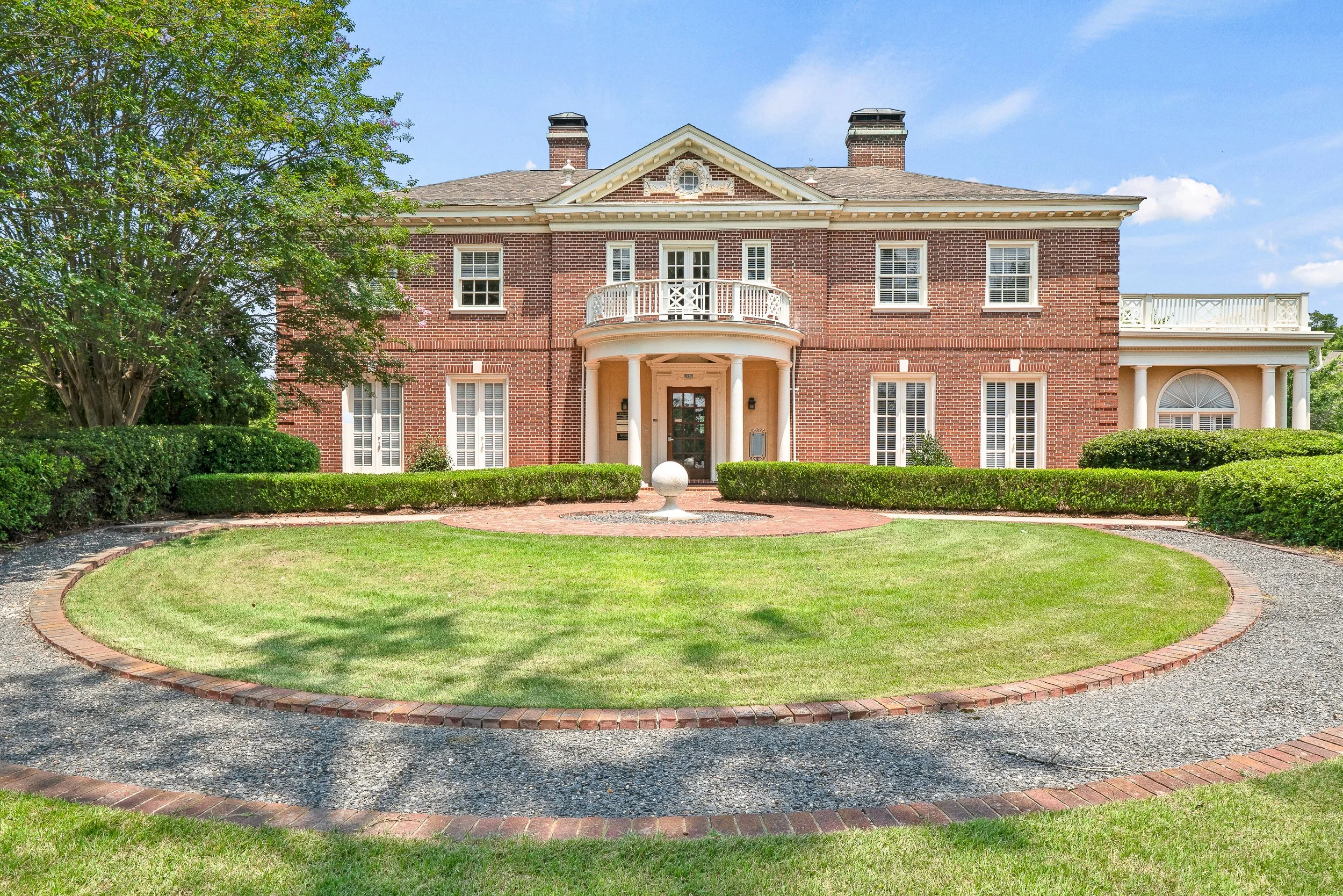 A large red brick house with white accents, multiple windows with shutters, a small balcony, and two chimneys. The house is surrounded by well trimming hedges, a lawn, and a curved gravel pathway with brick edging leading to the front door, under a blue sky with some clouds.