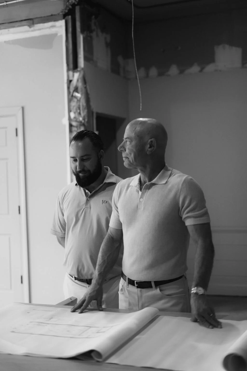 Two men, one senior with a bald head and the other younger with a beard, stand beside each other looking at blueprints on a table in a room under construction or renovation.