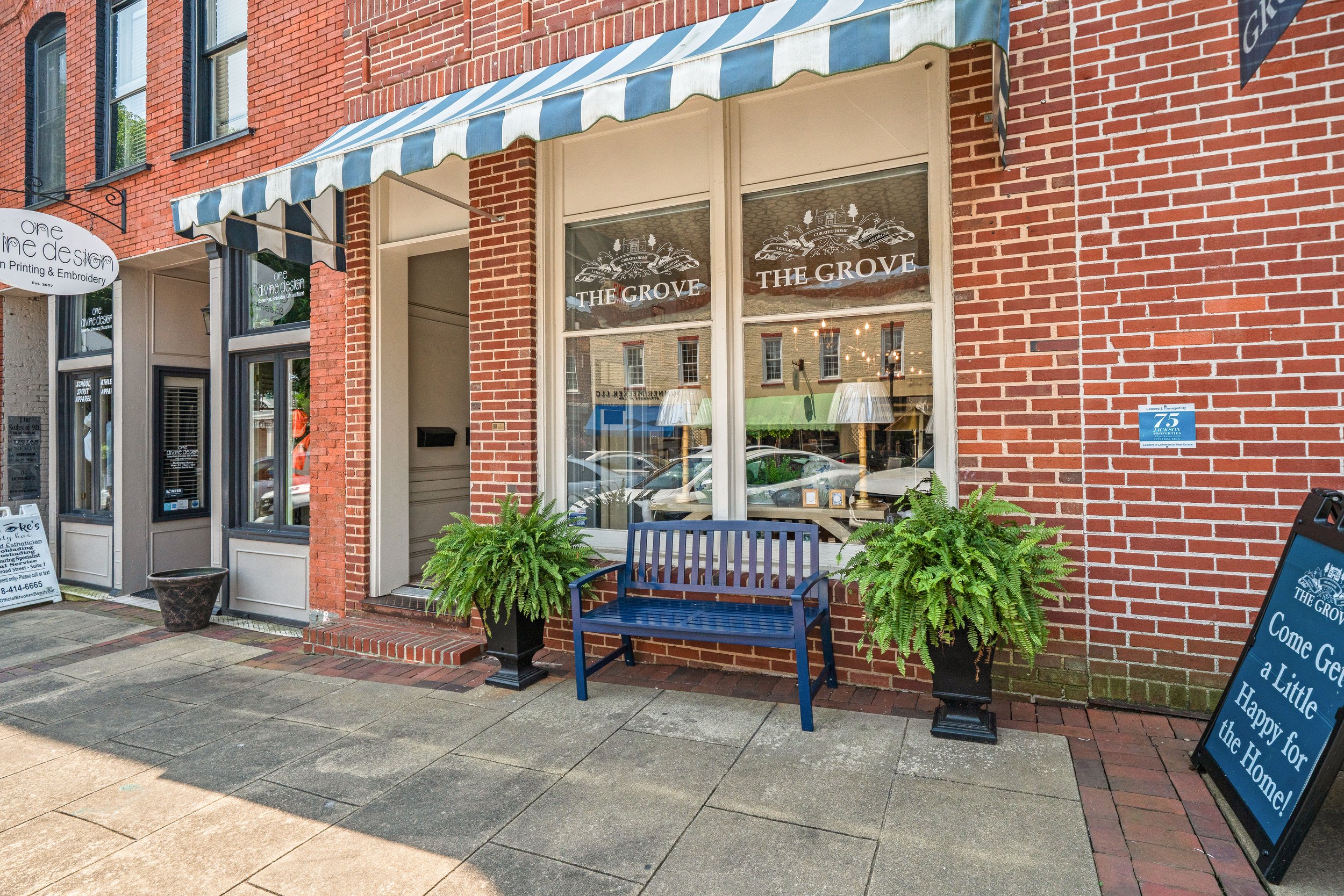 Storefront with a sign for 'The Grove' bakery or cafe, brick exterior, large window, blue and white striped awning, potted plants, a blue bench outside, and sidewalk signs.