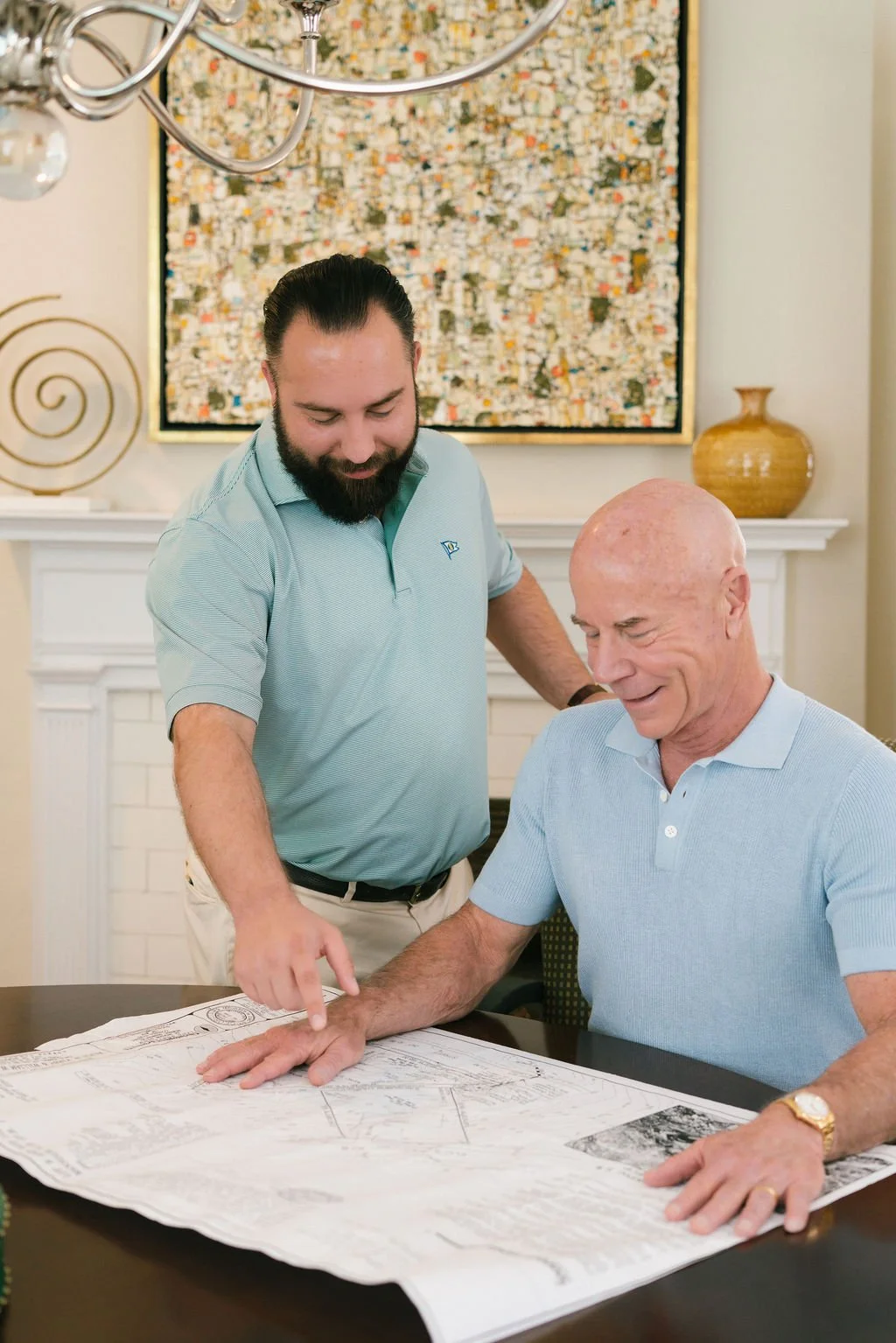 Two men look at a large paper plan or blueprint on a table, one pointing at it while the other smiles and looks on. They are in a room with a fireplace, artwork, and decorative items in the background.