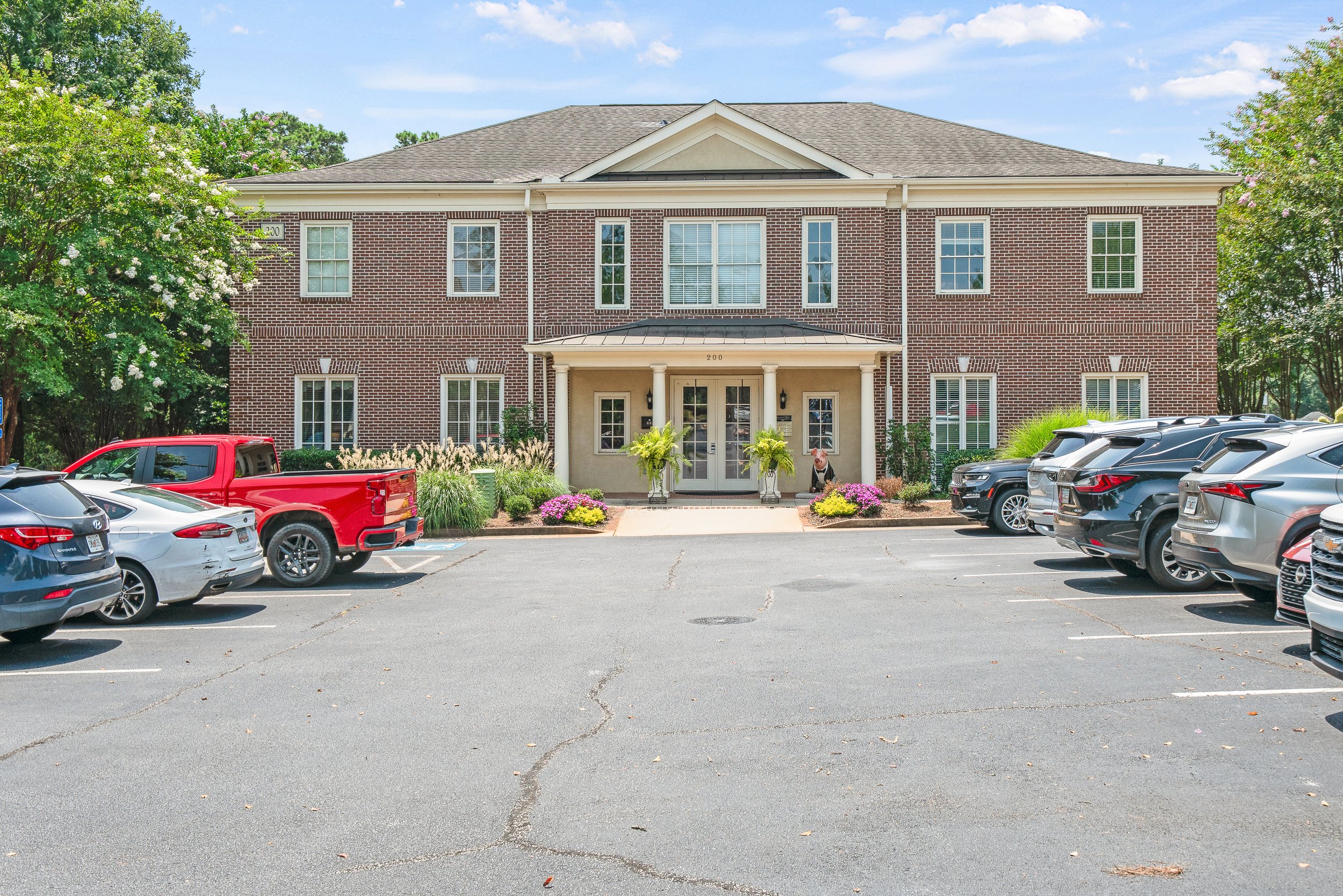 A brick apartment building with a covered entrance, surrounded by plants and flowers, and a parking lot filled with cars in the foreground.