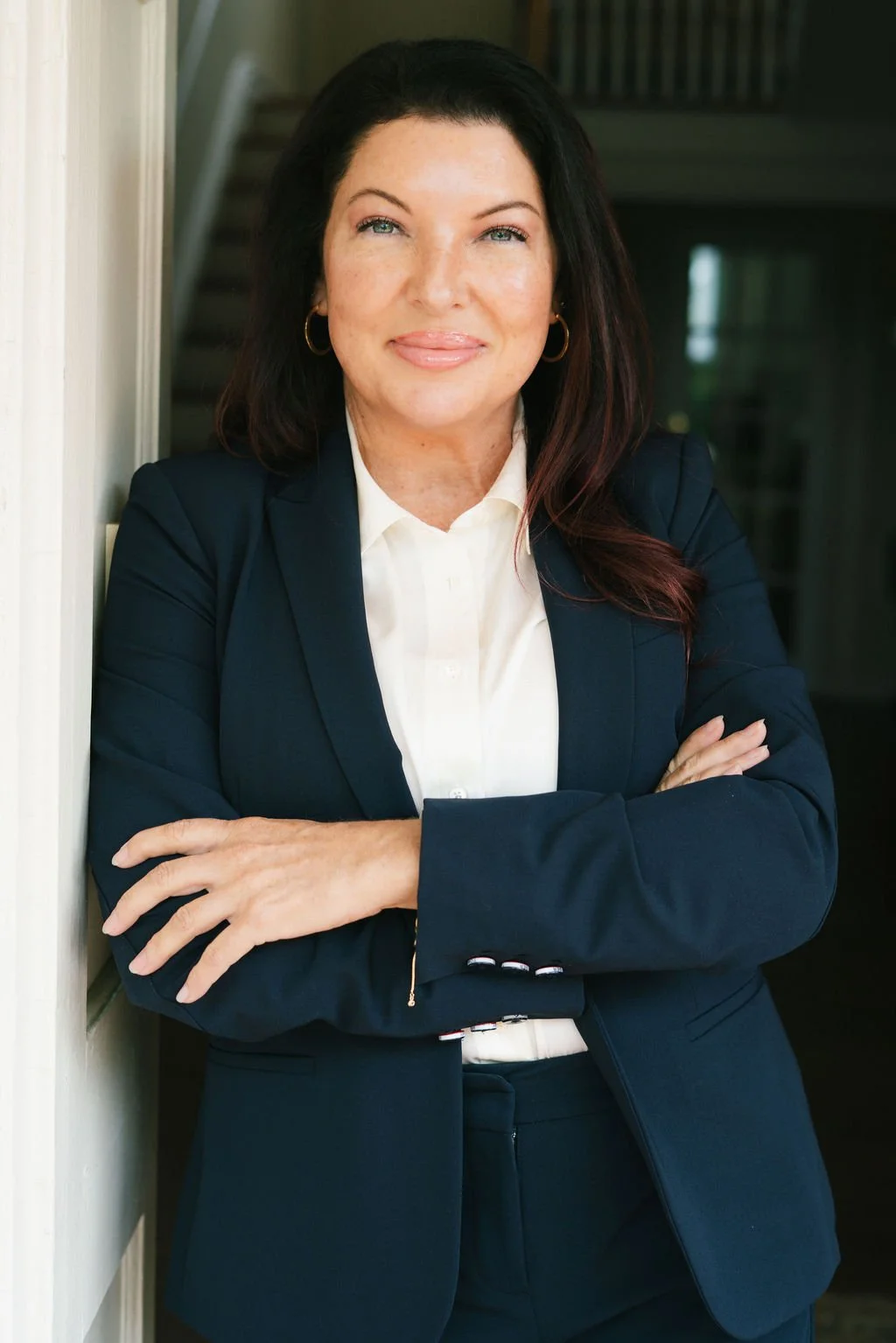 A woman with dark hair and fair skin wearing a navy blazer and white blouse is standing with arms crossed, smiling at the camera.