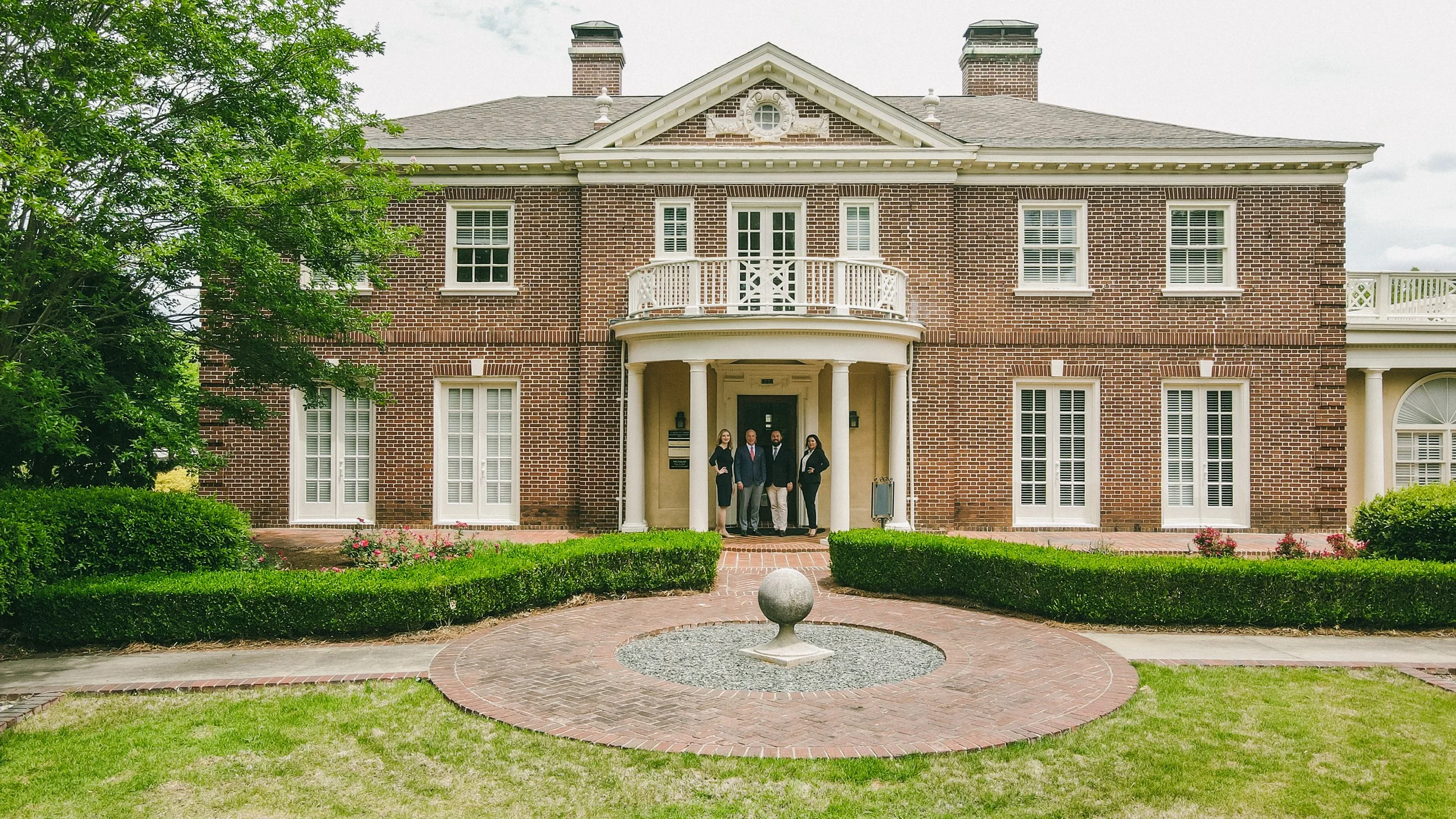 A large brick house with white windows and a covered front porch. Four people in formal attire are standing under the porch. The house has a small balcony above the porch and is surrounded by green lawn and bushes.