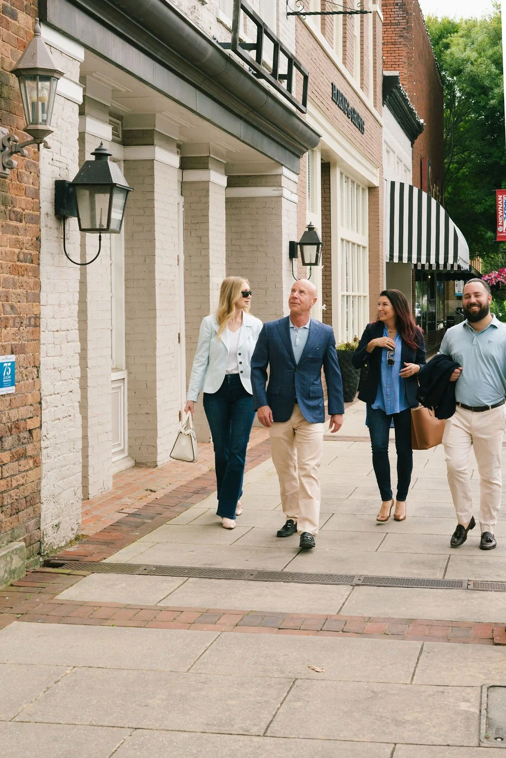 Four people walking on a city sidewalk, two women and two men, dressed in business casual attire, with brick and white buildings in the background.