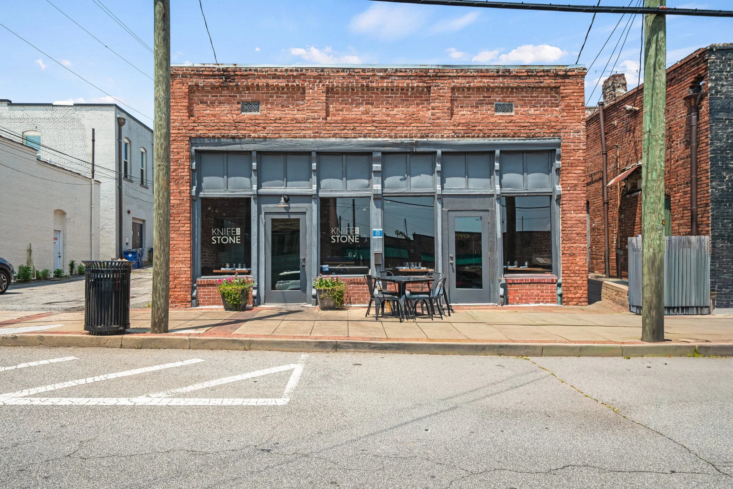 Front view of a brick building with large glass windows and patio furniture outside, in an urban setting with power lines and neighboring buildings.