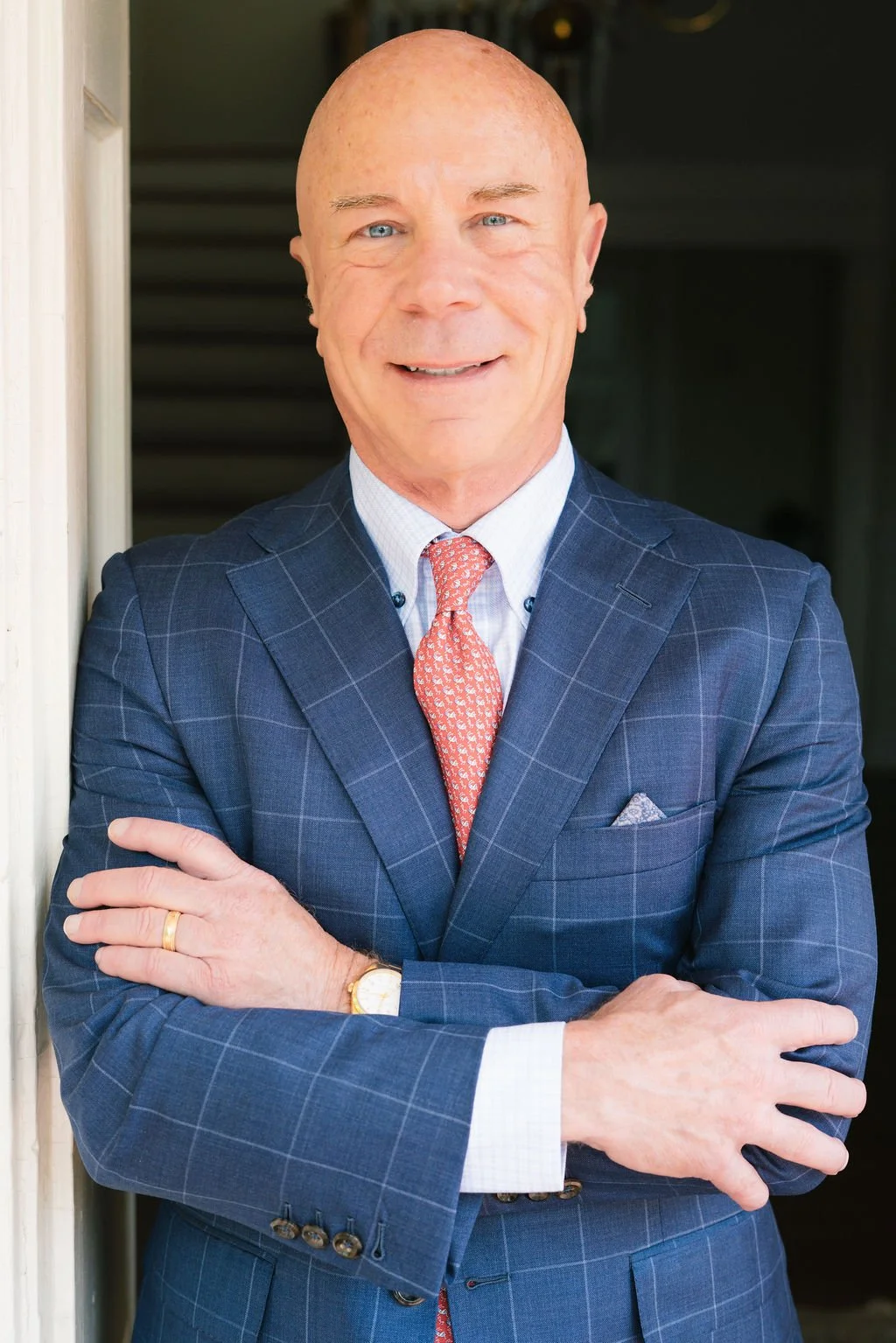 A bald man in a navy blue plaid suit with a white shirt and pink patterned tie, standing with arms crossed and smiling, in front of a dark background.