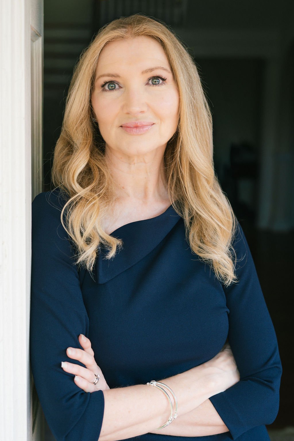 A woman with long blonde hair and blue eyes standing with her arms crossed, wearing a dark blue top, looking at the camera, with a neutral background.