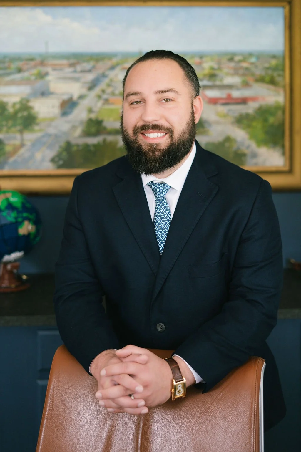 A man with a beard wearing a black suit, white shirt, and patterned blue tie, sitting and smiling at the camera. Behind him, there is a window with a cityscape view, and a globe is visible on a desk to the left.