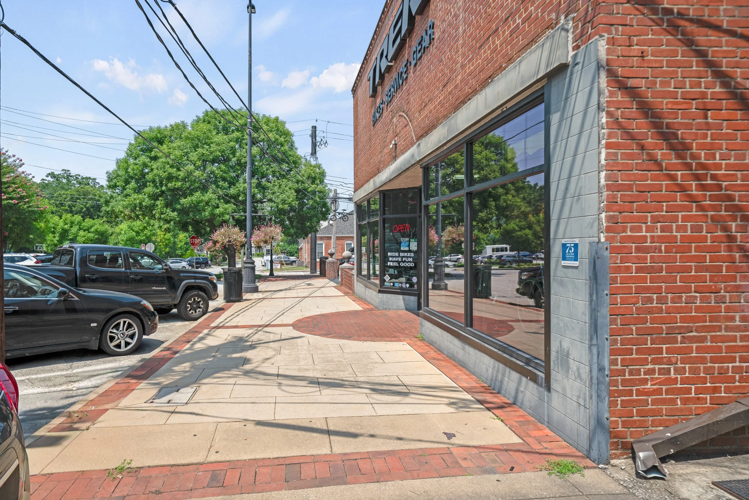 Street view of a brick building with a storefront, parked cars, a sidewalk, and trees under a partly cloudy sky.