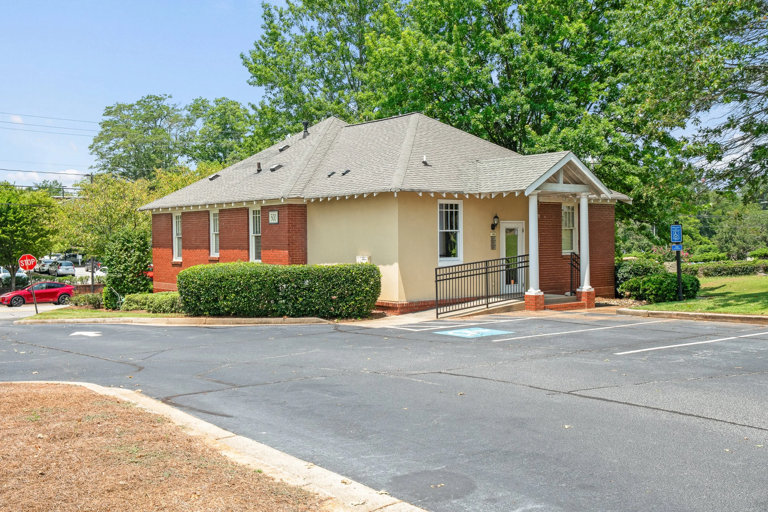 A small brick and beige building with a gabled roof, surrounded by green trees and a parking lot with marked spaces, including a handicapped spot.