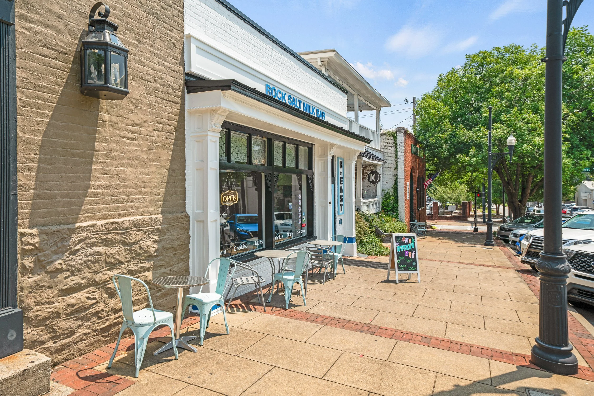 Exterior view of a small shop with a sign that reads 'ROCK SALT MILK BAR.' The shop has large front windows and outdoor seating with metal chairs and tables. The sidewalk is lined with lampposts and trees, under a clear blue sky.