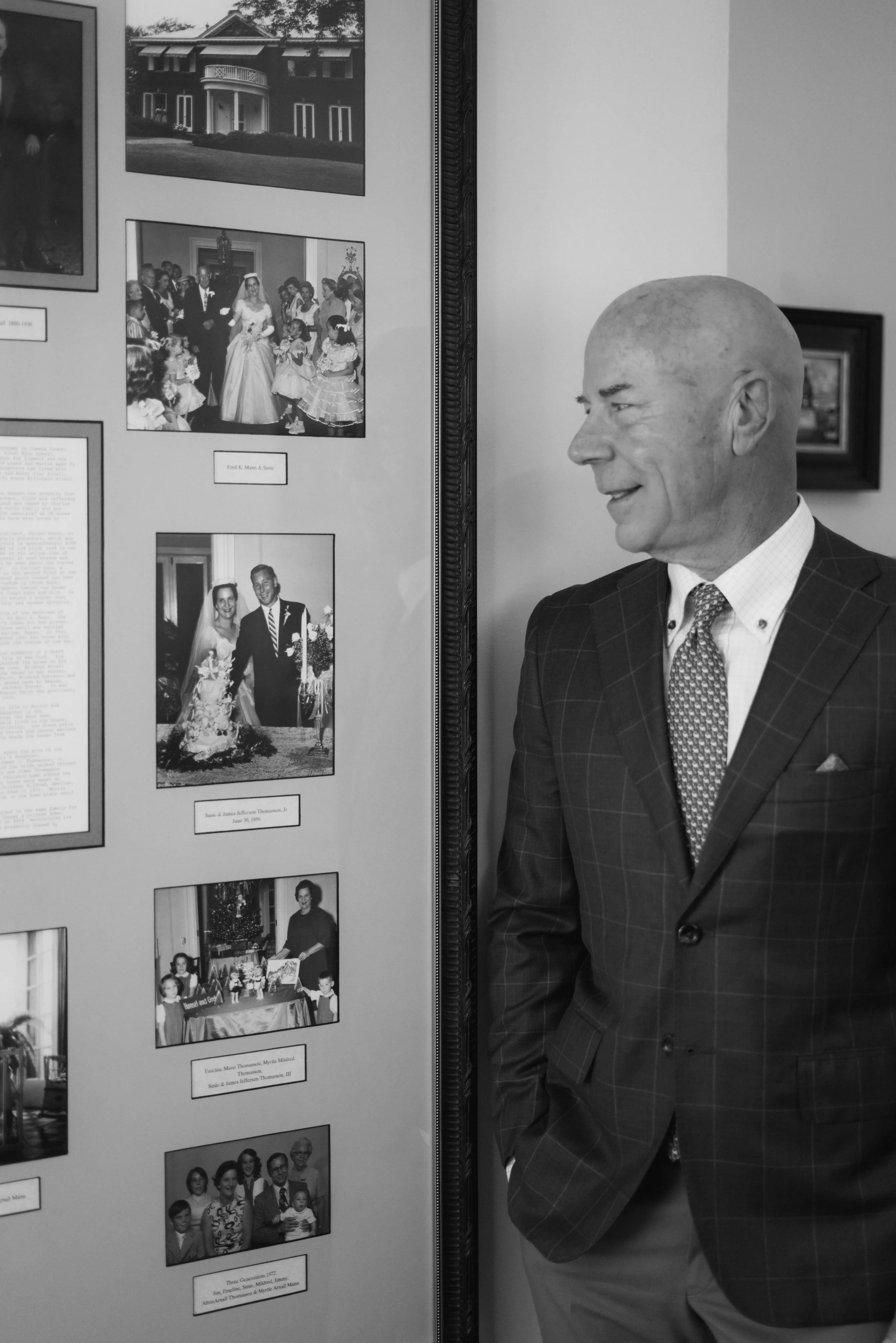 Black and white photo of a man in a suit smiling while looking at a display of framed family photos on a wall.
