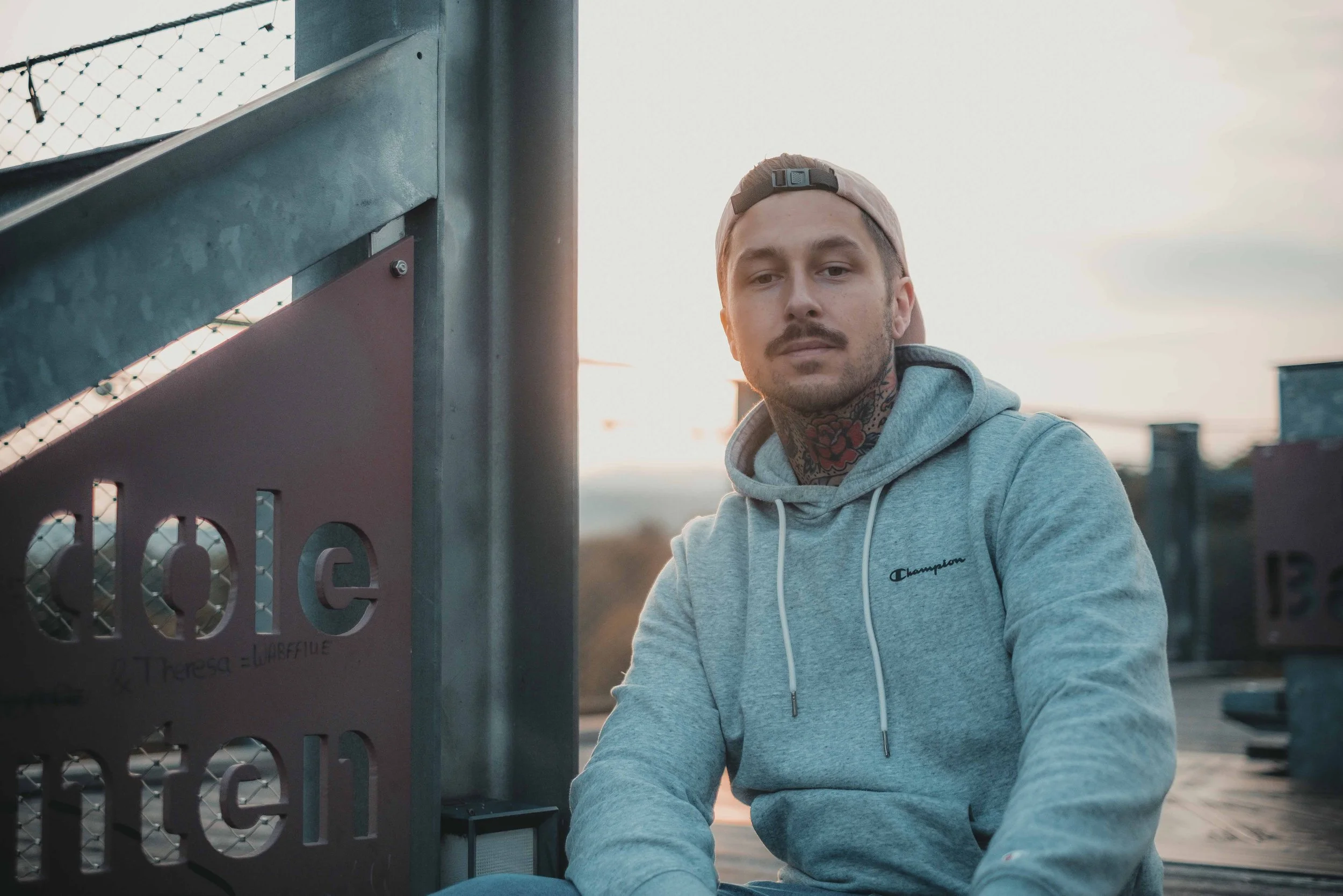 Ein Mann mit grauem Hoodie und Baseballkappe sitzt im Freien neben einem Metallzaun.