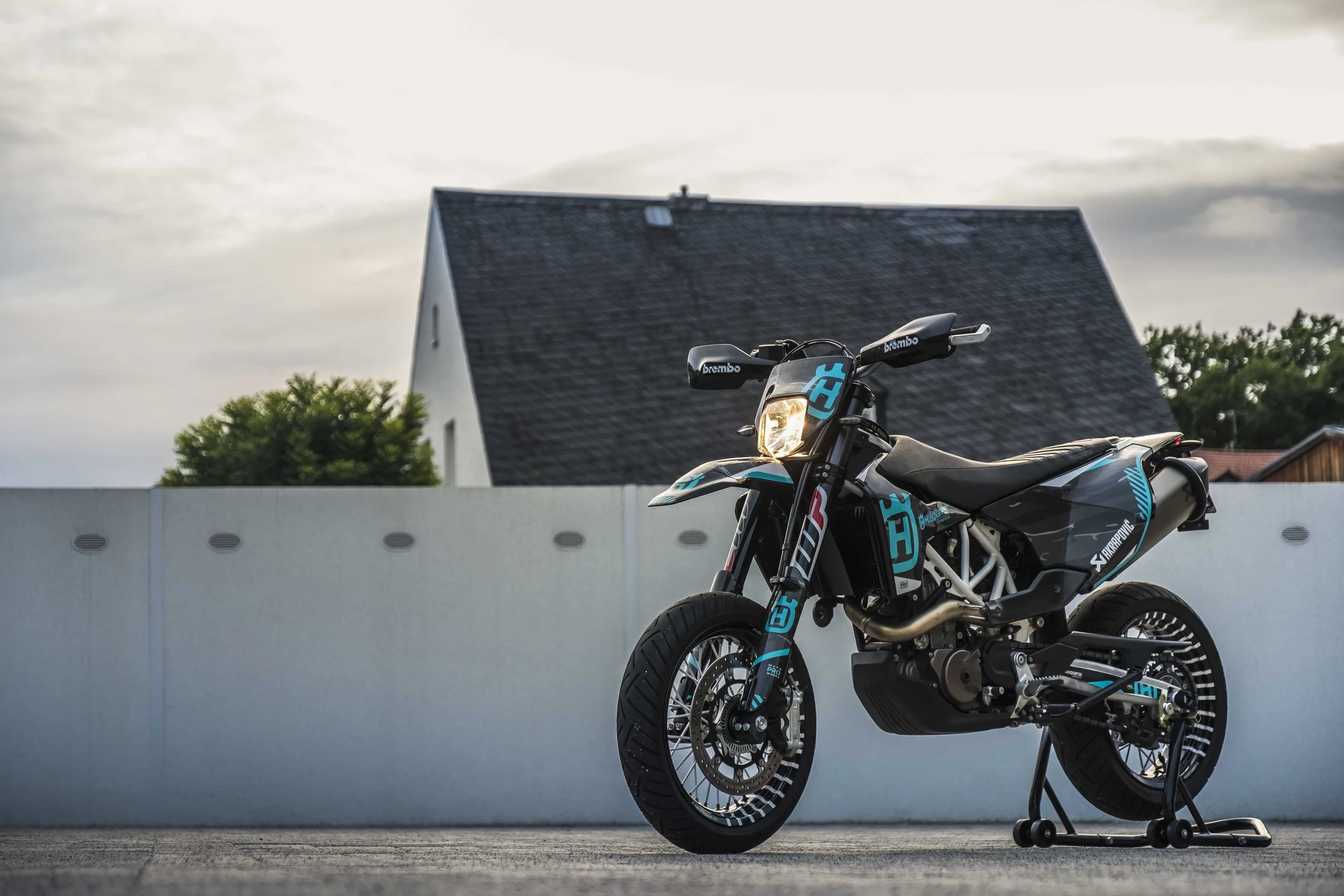 Ein schwarzes Supermoto-Motorrad mit blauen Akzenten steht vor einer weißen Mauer. Im Hintergrund ist ein Haus mit schiefergedecktem Dach sichtbar.