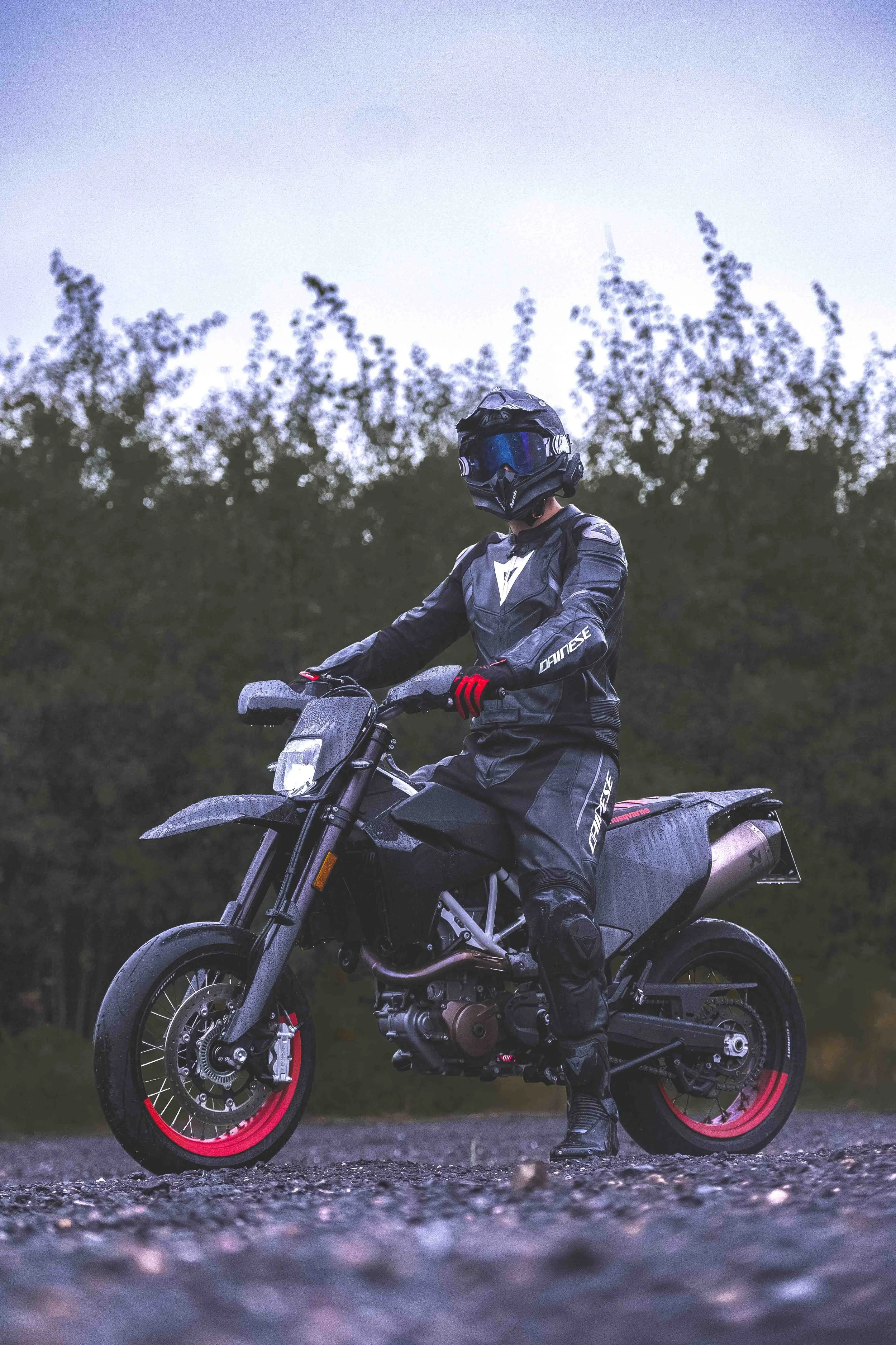 Motorradfahrer in schwarzer Schutzkleidung auf einer Straße vor Bäumen, sitzt auf schwarzem Motorrad mit roten Felgen.